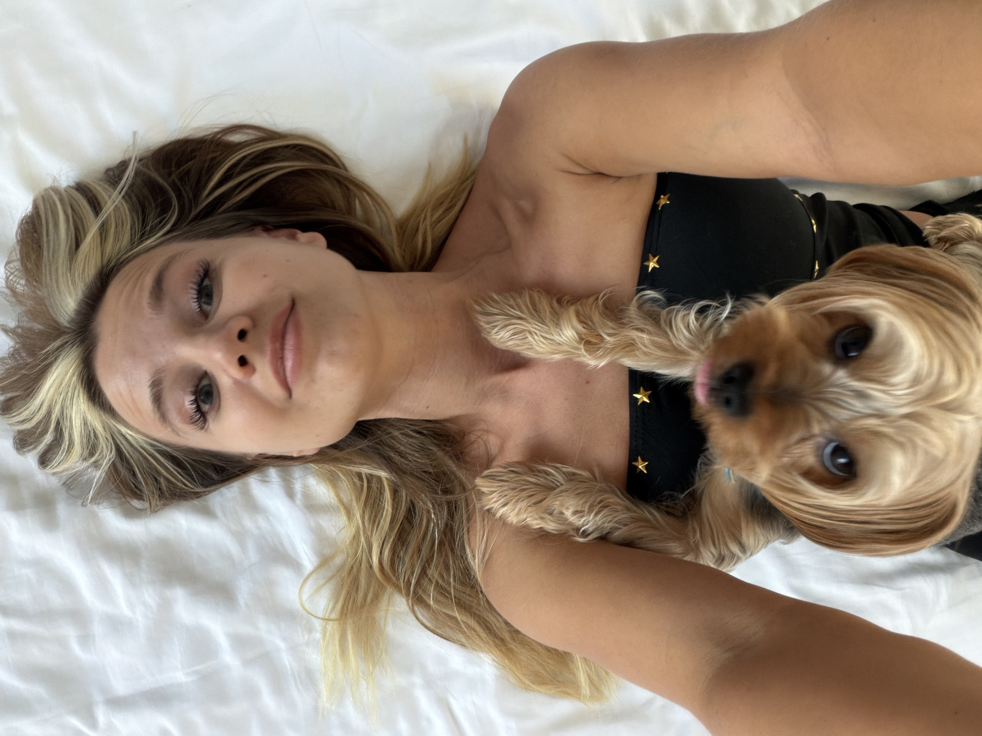 The media file features a close-up, overhead shot of a woman lying on a white surface, possibly a bed, with a small, light brown dog nestled beside her. The woman has blonde hair with lighter streaks and is looking towards the camera. She is wearing a black garment adorned with gold stars. The dog, a Yorkshire Terrier, is positioned in the foreground, with its face and paws visible. It appears to be looking up at the camera, with its tongue slightly out. The scene is lit by natural light, suggesting daytime, and the overall mood appears relaxed and intimate. The white surface the subjects are on creates a clean, uncluttered background, emphasizing the subjects. There is no visible text in the image. The location is identified as Thep Krasatti, Thailand.