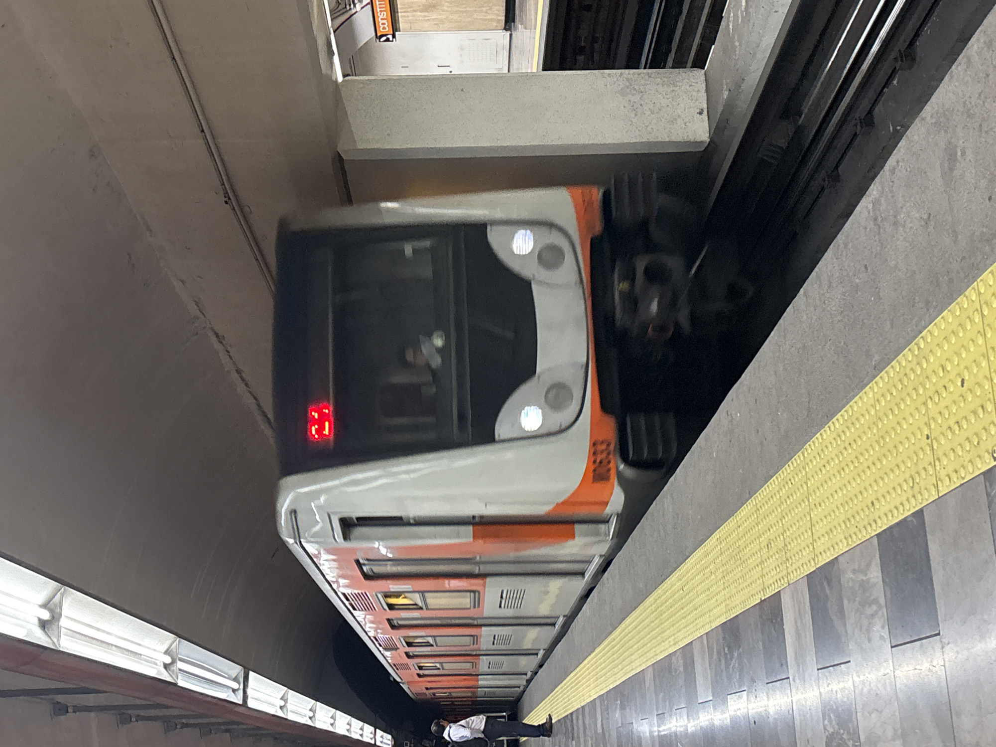 The image depicts a train arriving at a subway station in Mexico City, Mexico. The train is predominantly white and orange with the number "3" displayed in red LED digits on the front. The platform is grey with a yellow textured safety strip along the edge. A person wearing a white shirt and dark pants is lying on the platform. The environment is a subway station with overhead lights, concrete structures, and visible tracks.