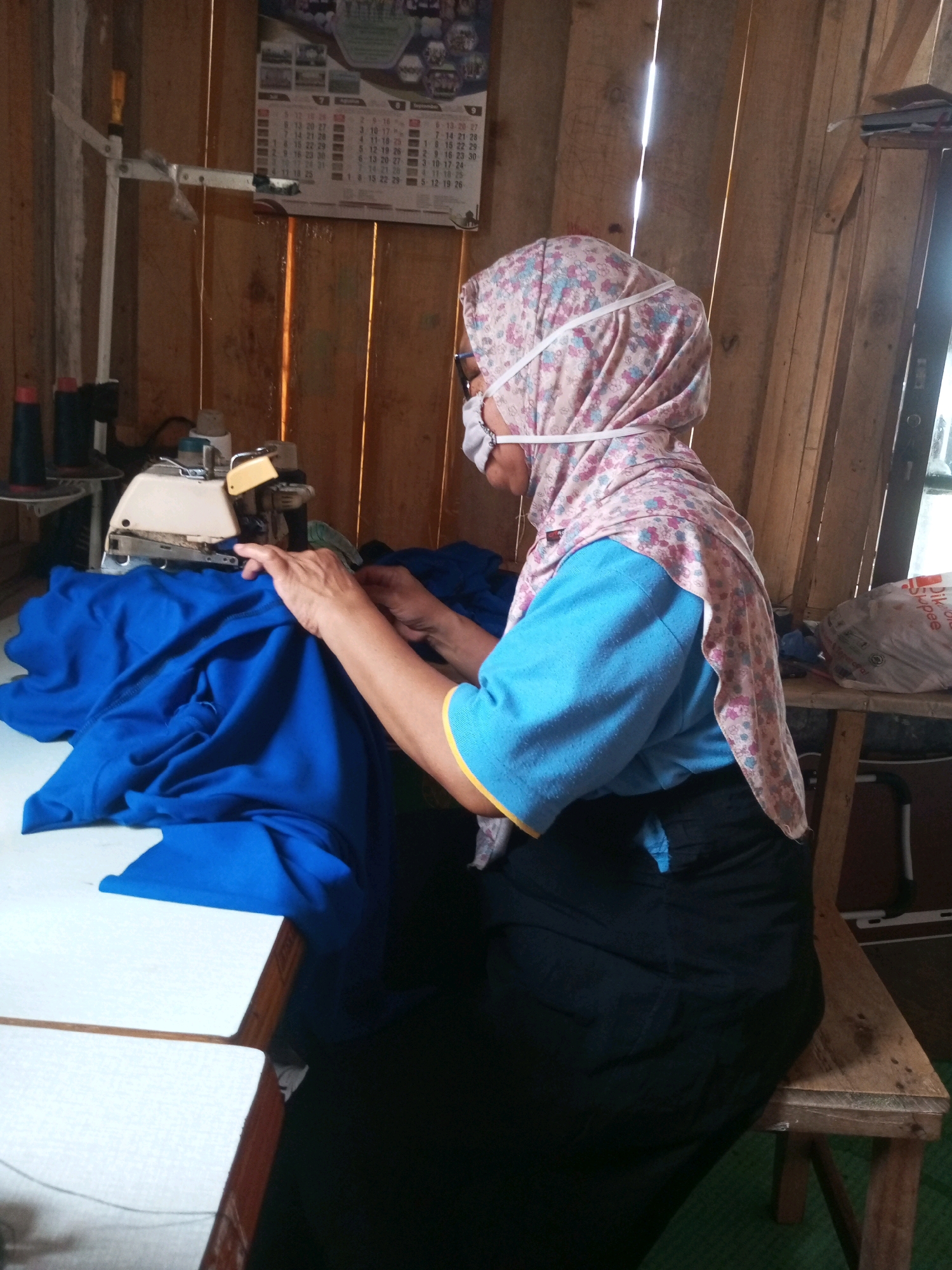 A woman, wearing a floral headscarf and a face mask, sits at a sewing machine, working on a bright blue fabric. She is positioned on the right side of the frame, facing left. The sewing machine is on a white table in the foreground. The background consists of wooden walls. A calendar is hanging on the wall behind the woman. The scene appears to be an indoor workspace in the village of Kalibeber, Indonesia, suggested by the presence of traditional wooden construction and items commonly found in such settings.