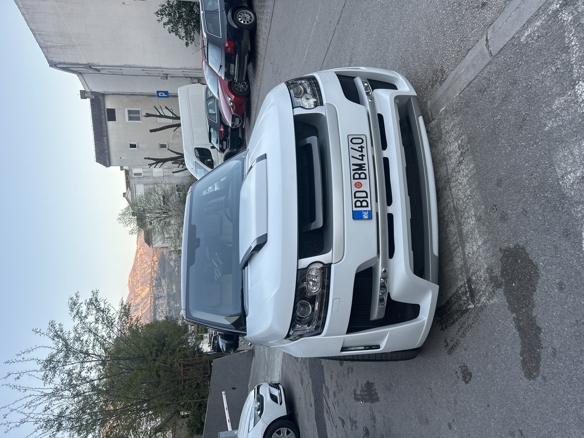 The image shows the front of a white Range Rover-style SUV parked on an asphalt street in Budva, Montenegro. The license plate reads "BD BM440". The vehicle is parked close to the curb. Behind the SUV, several other vehicles are visible, including a white van, a red car, and a dark-colored car. A multi-story building is partially visible in the background on the left side, as well as the mountains. A tree is also present in the lower left of the frame. The asphalt shows some staining and markings.