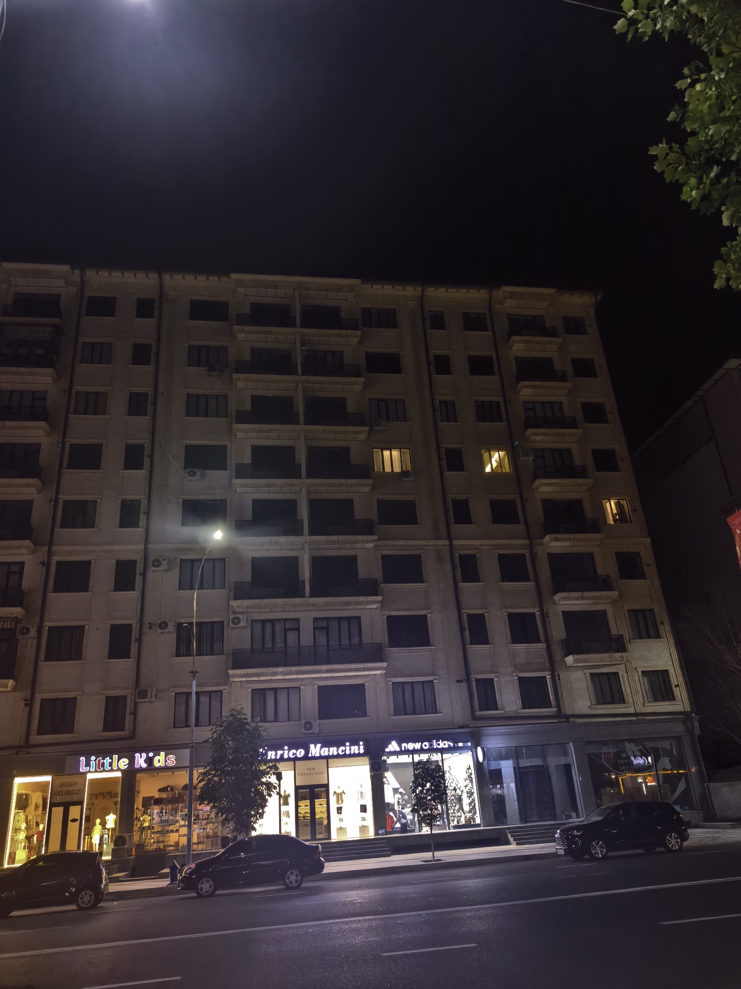 A night-time, wide-angle, eye-level shot captures the facade of a multi-story building in Fergana City, Uzbekistan. The building, likely an apartment complex with retail spaces on the ground floor, is illuminated by streetlights and interior lights from the shops. The upper floors feature numerous dark windows and balconies. The ground floor retail spaces are brightly lit, displaying merchandise. Visible storefronts include "Little K'ds," "Enrico Mancini," and "New Adidas." Two dark-colored cars are parked on the street in front of the building, and the street itself is visible in the foreground, marked with white lines. A single streetlamp casts a bright glow. Trees are visible to the right of the building and in front of the "New Adidas" store. The sky above is dark, with a faint light source visible towards the top left.
