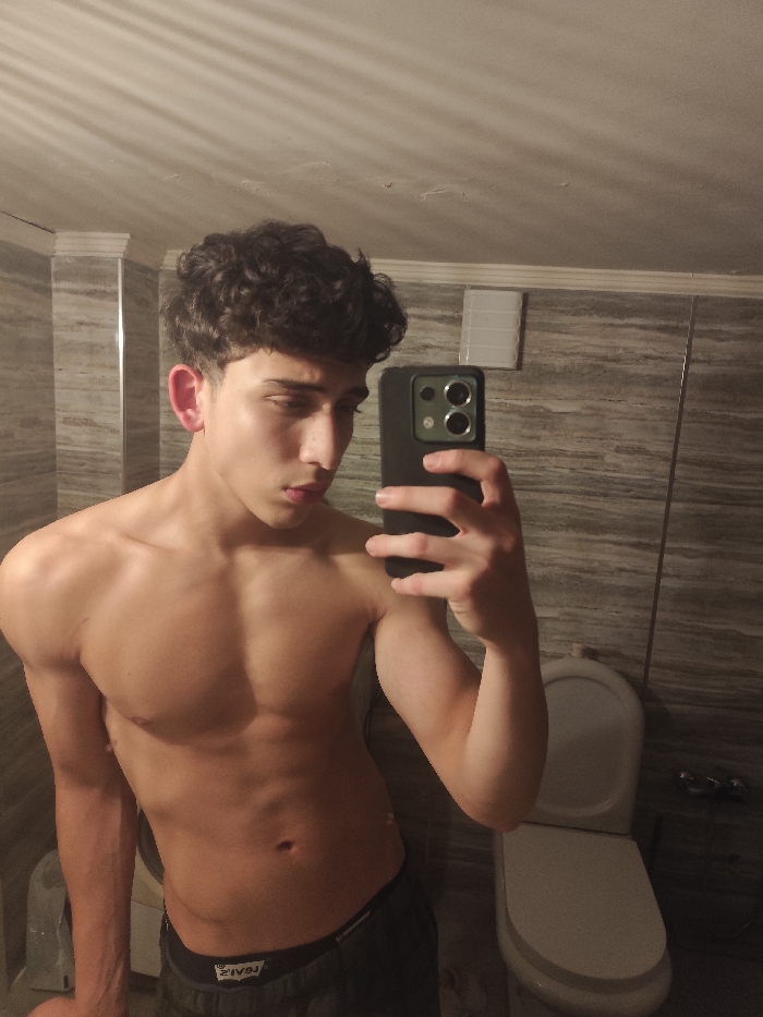 A shirtless young man with curly hair takes a selfie in a bathroom. He holds a dark-colored smartphone to capture the image, tilting his head slightly downward. The bathroom features gray-toned tiled walls, a toilet, and visible overhead lighting casting shadows. His physique reveals defined pectoral and bicep muscles. He wears dark-colored bottoms.