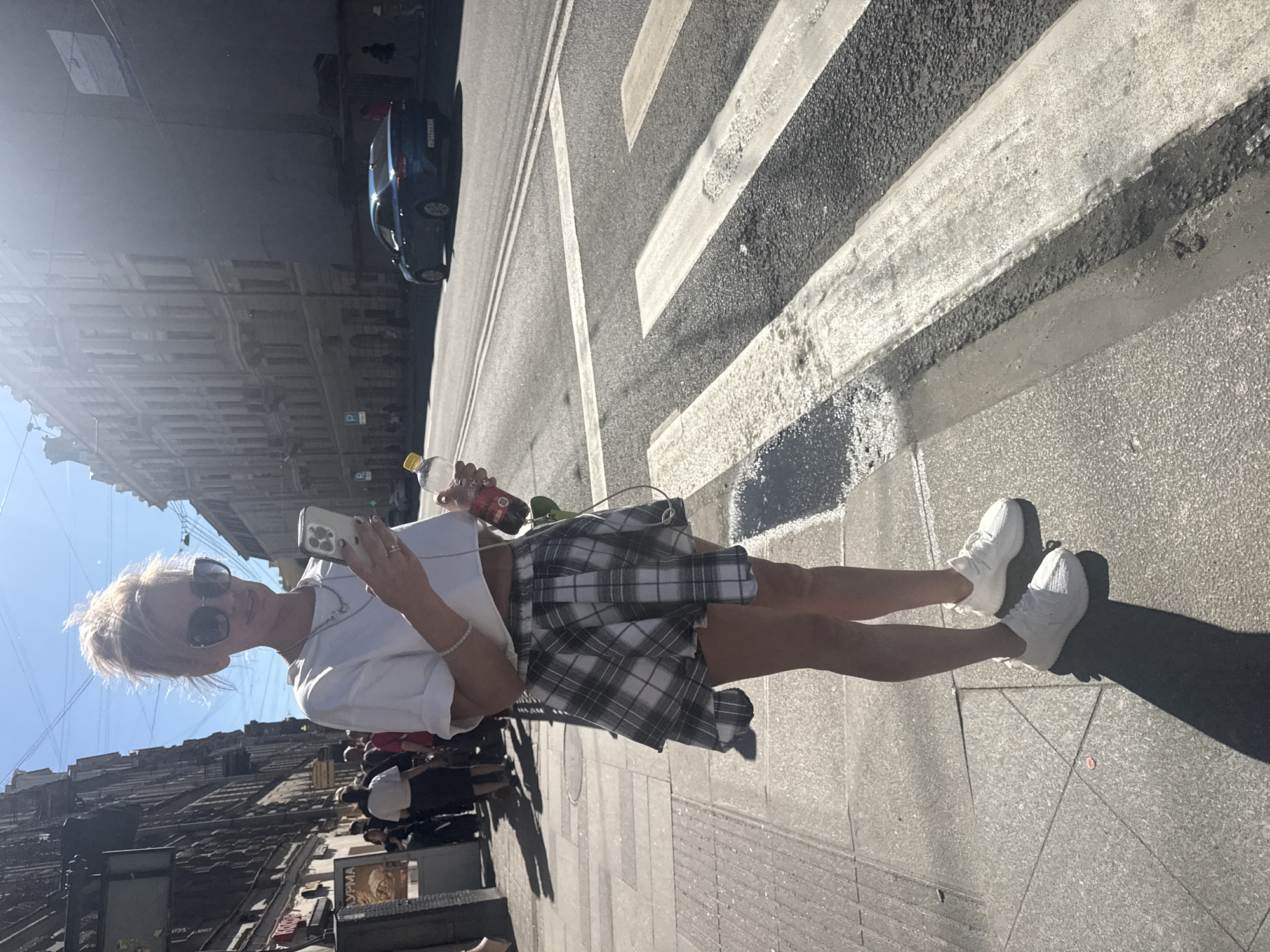 A young woman stands on a sidewalk in Saint Petersburg, Russia, looking at her phone. She has blonde hair pulled up in a messy bun and is wearing oversized sunglasses, a white crop top, and a plaid mini-skirt. She is holding a plastic water bottle in her right hand. Behind her, pedestrians walk along the sidewalk next to a street with traffic. The architecture appears to be historic, with multi-story buildings lining the street. A blue car is visible driving on the road. A pedestrian crosswalk is in the foreground.