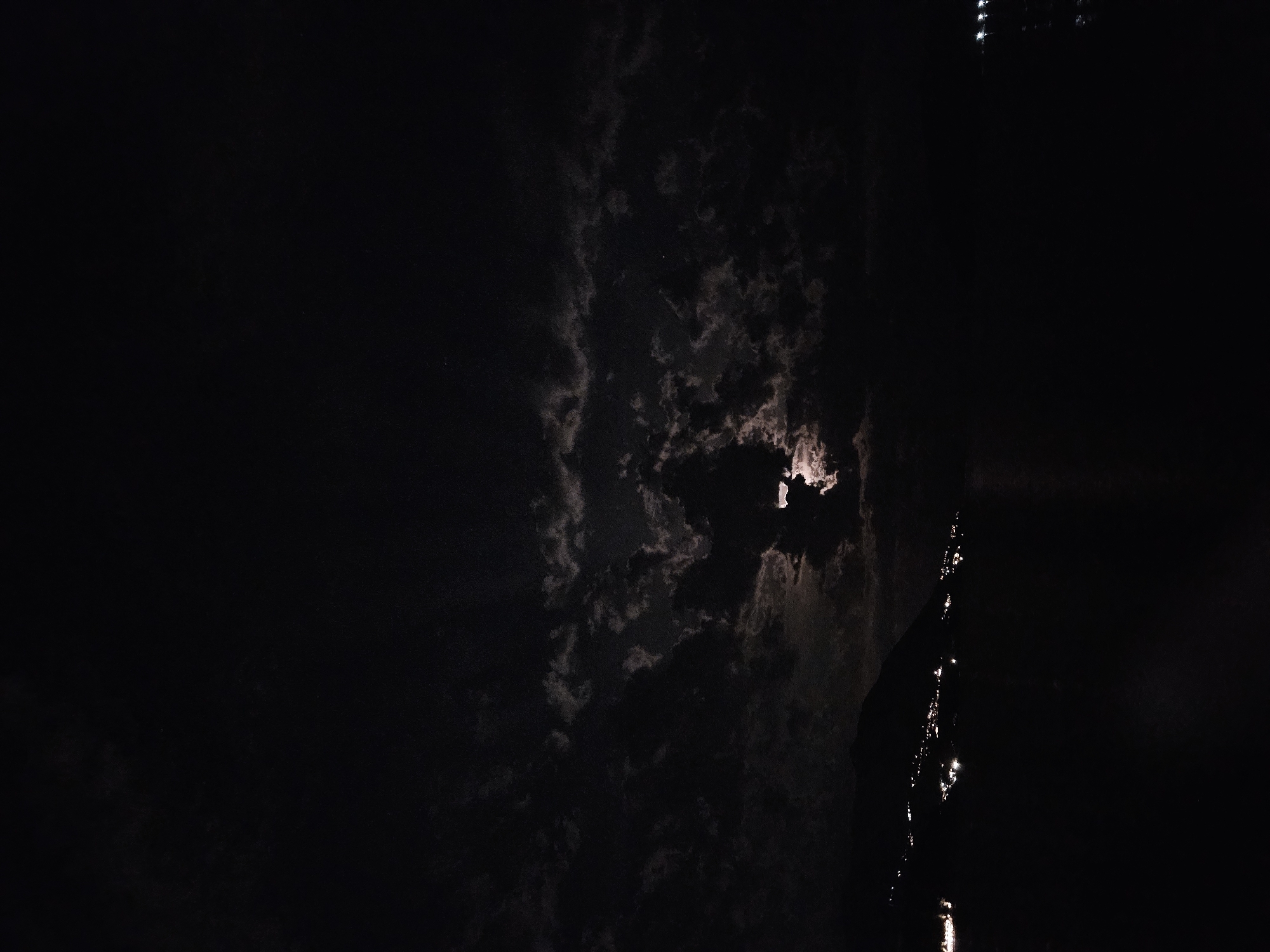 This vertically oriented image captures a dramatic night scene, likely over a body of water, under a cloudy sky. The dominant feature is a bright, partially obscured light source, strongly resembling the moon, positioned in the upper-right portion of the frame. It pierces through thick, dark clouds, casting an ethereal glow and illuminating their edges with shades of white and grey against the otherwise black sky.
On the far right side of the image, a dark, indistinct silhouette of land or a large structure runs vertically from top to bottom. Below this silhouette, a body of water is visible, reflecting elongated streaks of light. These reflections suggest distant light sources along the unseen shoreline or the moon's light dancing on gentle ripples. The left and top-left areas of the image consist of vast expanses of dark sky, with hints of very subtle cloud texture.
The overall impression is one of a serene yet mysterious night, characterized by the interplay of deep shadows and soft, radiant moonlight through a heavily clouded sky. There is no visible text.
