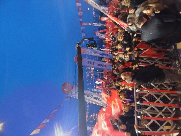 The image shows a large crowd of people gathered in an outdoor space in Istanbul, Türkiye. The location of the crowd is near the coordinates 41.084, 28.678, which corresponds to a street near Taksim Square. The crowd is standing behind a metal barrier and is holding red flags with white text that says "CHP" and "CHP SILE". The crowd is facing a stage that is not visible in the image. In the foreground of the image are a large number of people wearing jackets, coats, and hats. The crowd is diverse in age and gender, with many families present. The crowd appears to be waiting for an event to begin. The sky in the image is blue with some clouds. The event is likely a political rally or a campaign stop for the CHP (Republican People's Party) political party.