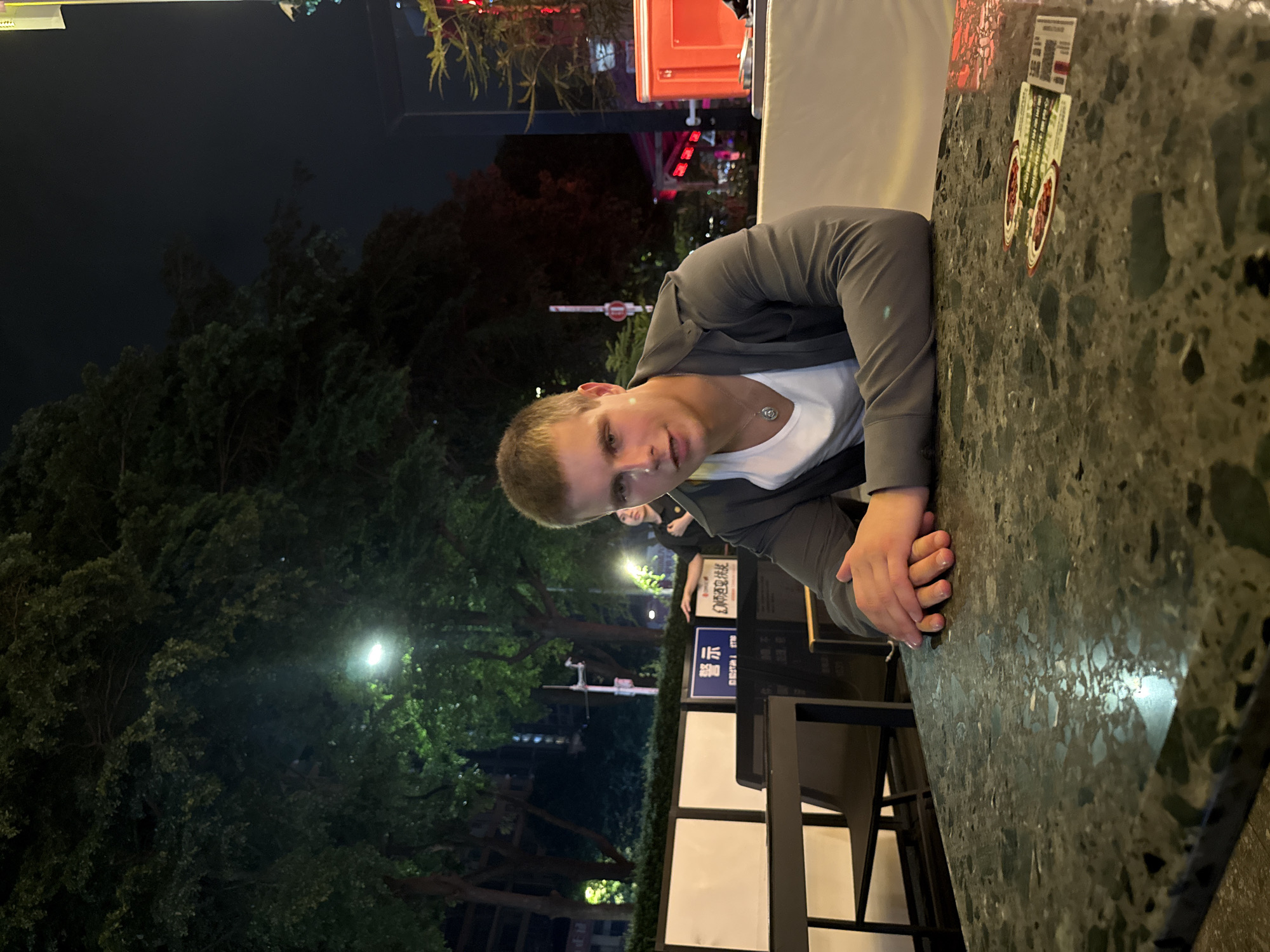 A young man is seated at an outdoor table in what appears to be the Tianhe District of China. The table has a dark, speckled surface with stickers attached to it. The man has short blond hair and is wearing a grey jacket over a white t-shirt. He is resting his arms on the table. Behind him, there are trees and streetlights, with Chinese signage visible on a barrier. In the background, there is a glimpse of a lighted structure and dark sky. A chair is seen positioned near the table.