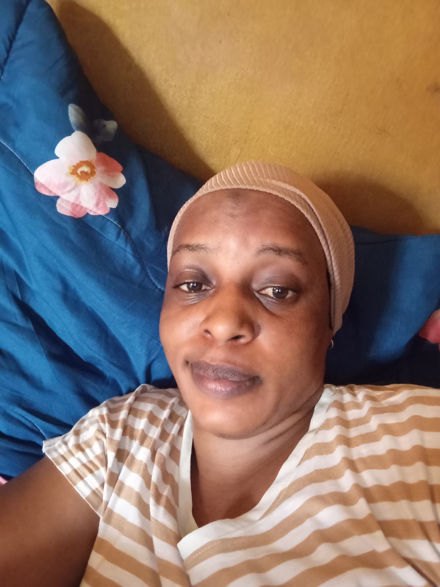 The image depicts a person with dark skin, wearing a beige ribbed head covering and a tan and white striped shirt. The person is lying on a blue fabric with a pink and white flower design. Behind the fabric is a yellow surface. The location is specified as Narayi, Nigeria, but no evidence in the image supports the location specification. No actions or interactions are depicted.