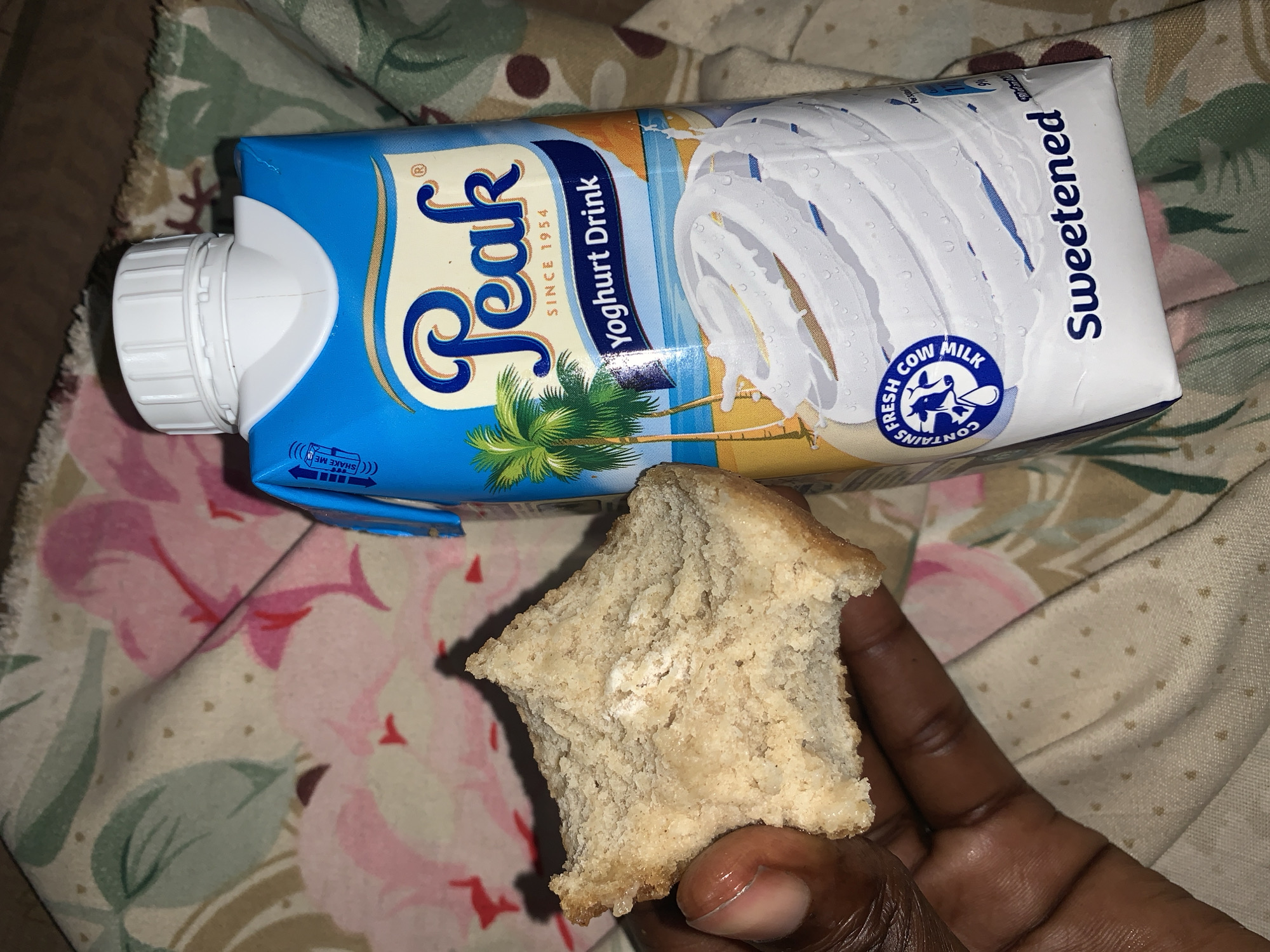 The image displays a close-up of a person's hand holding a piece of bread or cake, placed on a patterned fabric, alongside a carton of yogurt drink.
**Subjects:** The main subjects are a dark-skinned hand holding a food item and a packaged "Peak" brand yogurt drink.
**Objects:** The yogurt drink carton is predominantly blue and white, featuring the brand name "Peak" and the text "Yoghurt Drink" and "Sweetened." It states "SINCE 1954" and includes a circular emblem that reads "CONTAINS FRESH COW MILK" with a cow illustration. An icon with arrows and "SHAKE ME" is also visible. The carton has a white screw cap and shows an illustration of a palm tree and a beach scene. The food item is an irregularly shaped piece of light brown bread or cake with a somewhat coarse texture. The background is a fabric with a floral pattern in pink, green, and beige tones.
**Activity:** The activity appears to be snacking or preparing to eat, with a drink accompanying the food.
**Notable details:** The scene suggests a casual, indoor setting, possibly a home in Rafin Par, Nigeria. No specific time of day or emotions are discernible.
**Visible text:** "CONTAINS FRESH COW MILK", "SHAKE ME", "Peak", "SINCE 1954", "Yoghurt Drink", "Sweetened".