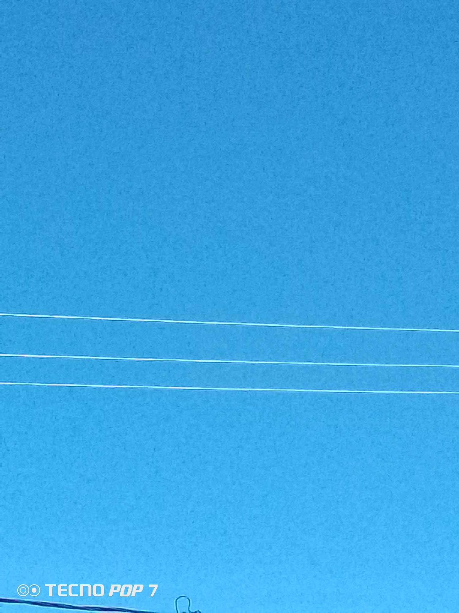 The image displays a clear, bright blue sky with minimal cloud cover, situated in Altagracia, Venezuela. Three parallel, thin white lines, consistent with utility wires, extend horizontally across the mid-to-lower section of the frame. A faint, thinner white line is also barely visible along the upper edge of the image. In the bottom-left corner, a white digital watermark is present, featuring a circular camera-like icon followed by the text "TECNO POP 7". A dark, indistinct, curved object is partially visible at the very bottom-right corner of the image. No people or specific actions are depicted.