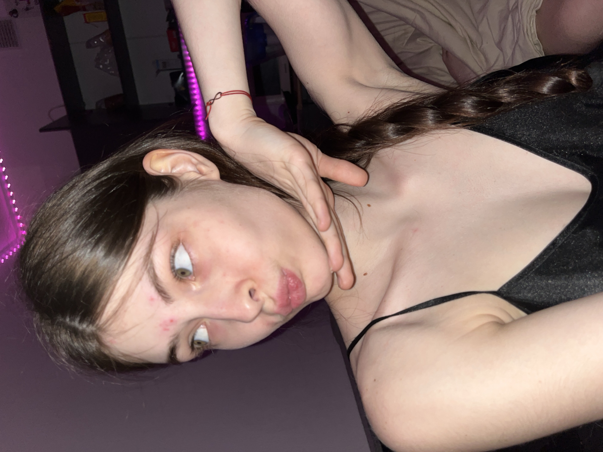 A close-up shot shows a young woman with light skin, brown hair, and green eyes lying down. She wears a black top with thin straps. Her head is tilted, and her face displays a slight pout. Another person's hand, also with light skin, is positioned near her neck. A red bracelet adorns the wrist of this hand. The background includes hints of a room with purple lighting and furniture. There is no evidence indicating the location is Saint Petersburg, Russia.