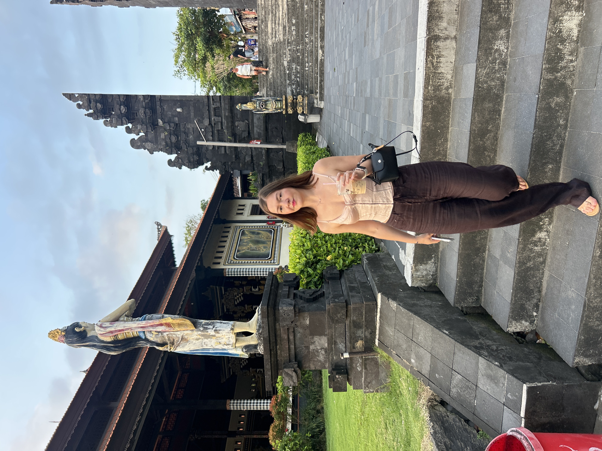 A woman is descending a stone staircase outdoors, holding a drink and a mobile phone. She is wearing a pale pink corset-style top and dark brown wide-leg pants, with light-colored sandals. A black crossbody bag is slung over her shoulder. Behind her, to the left, is a large stone statue of a woman in traditional attire, positioned in front of a building with a dark tiled roof. To the right, the staircase leads up towards ornate, dark stone structures, possibly part of a temple or gate. Green grass and lush bushes are visible on the left side of the image. In the background, on the stairs to the right, two other individuals are walking upwards. The sky above is partly cloudy. This scene appears to be at the Pura Mandara Giri Baloongan in Kediri, East Java, Indonesia, known for its distinctive statues and architectural style.