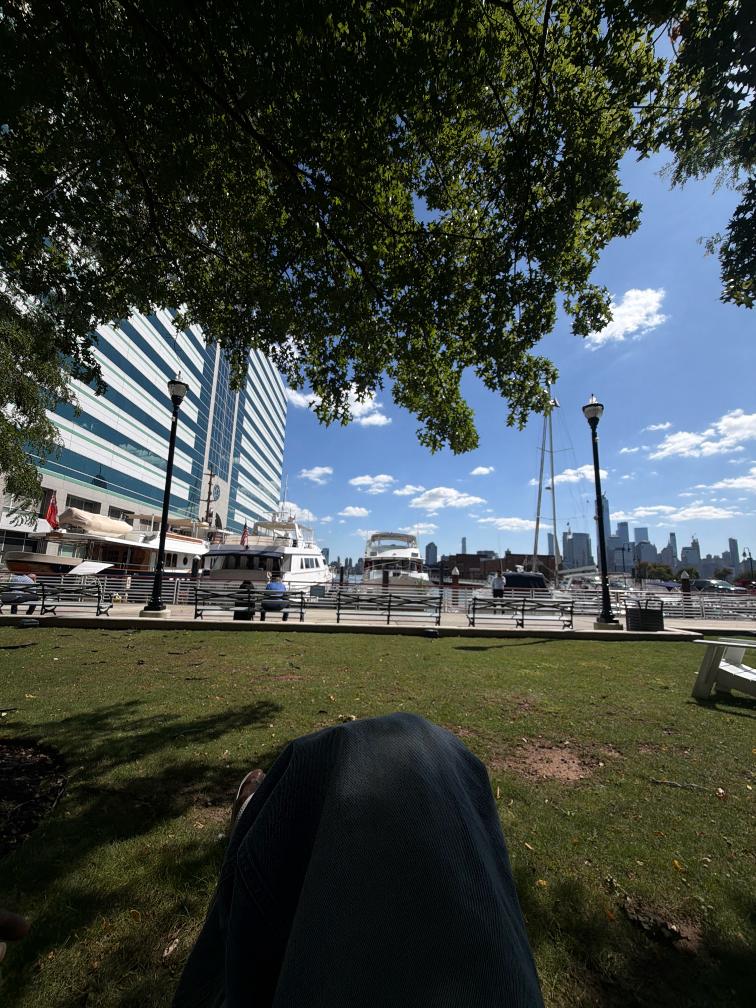 In Jersey City, USA, under a canopy of green leaves and a bright blue sky dotted with white clouds, a park scene unfolds. A person, whose legs and torso are visible in the foreground, is seated on the grass. The individual is wearing dark-colored pants.
Beyond the park, a waterfront promenade is lined with benches. A lamppost stands prominently on the promenade. Several large yachts are docked along the waterfront, with some displaying American flags. Behind the yachts and promenade, a tall, modern building with a white and blue striped facade dominates the middle ground. In the distance, a city skyline, characterized by numerous skyscrapers, stretches across the horizon. The overall environment suggests a clear, sunny day at a waterfront urban park.
