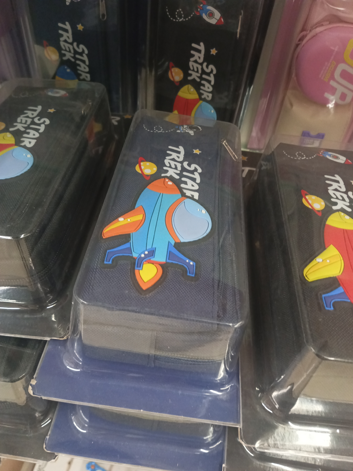 In Peshawar City Tehsil, Pakistan, a retail display features several Star Trek-themed pencil cases. These cases are dark blue with colorful cartoon graphics of rockets, planets, and astronauts. The items are packaged in clear plastic. The lighting suggests an indoor environment, likely a store.