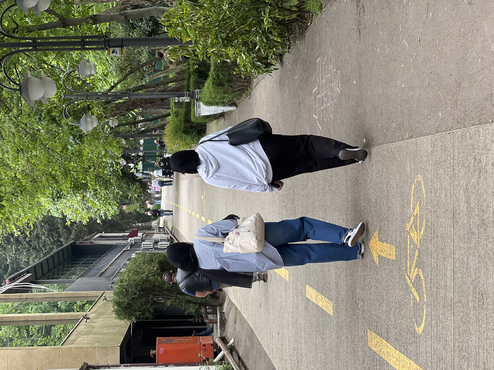 The image captures a sidewalk scene in Tbilisi, Georgia. Two women, both wearing head coverings, are walking in the same direction on the sidewalk. One is wearing a light-colored hoodie and black pants, while the other is wearing a striped shirt and jeans. The sidewalk features yellow painted markings, including directional arrows and a bicycle symbol. The background includes trees, lamp posts, and buildings. Other pedestrians are seen further down the sidewalk.
