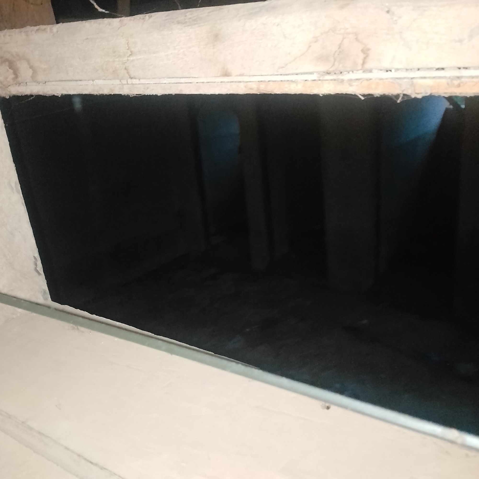 The image depicts a dark, unfinished interior space, possibly a crawl space or a room under construction. The visible area shows rough, unpainted walls and what appears to be exposed insulation along the top edge of an opening. The floor is uneven and dark, with some debris visible. The lighting is dim, casting long shadows and obscuring details within the space. The location in Ogbomosho, Nigeria, is not visually evident from the provided image.