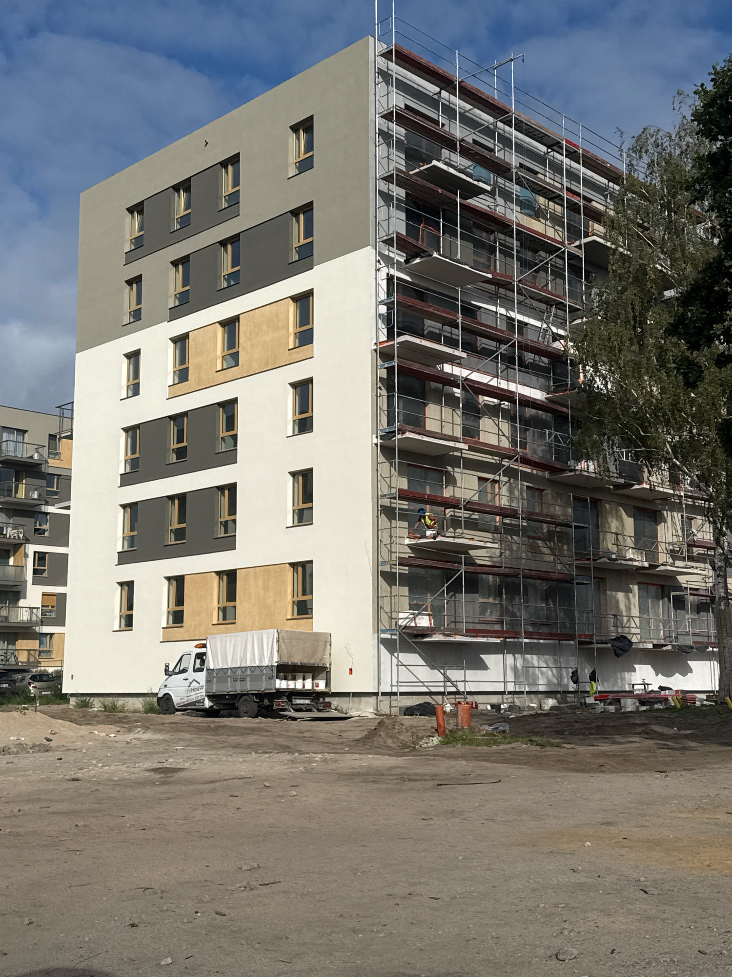 The image captures a construction site in Nowa Sól, Poland, focused on a multi-story apartment building undergoing exterior work. The building features a modern design with sections of light grey, dark grey, and cream-colored facades, punctuated by regularly spaced windows with wooden frames. Scaffolding completely envelops the right side of the building, indicating ongoing renovation or construction. A white truck with a covered cargo bed is parked near the building's entrance on the left side, and several construction barrels and materials are visible on the ground. The environment is a mix of unfinished urban landscape and greenery from trees to the right of the building.