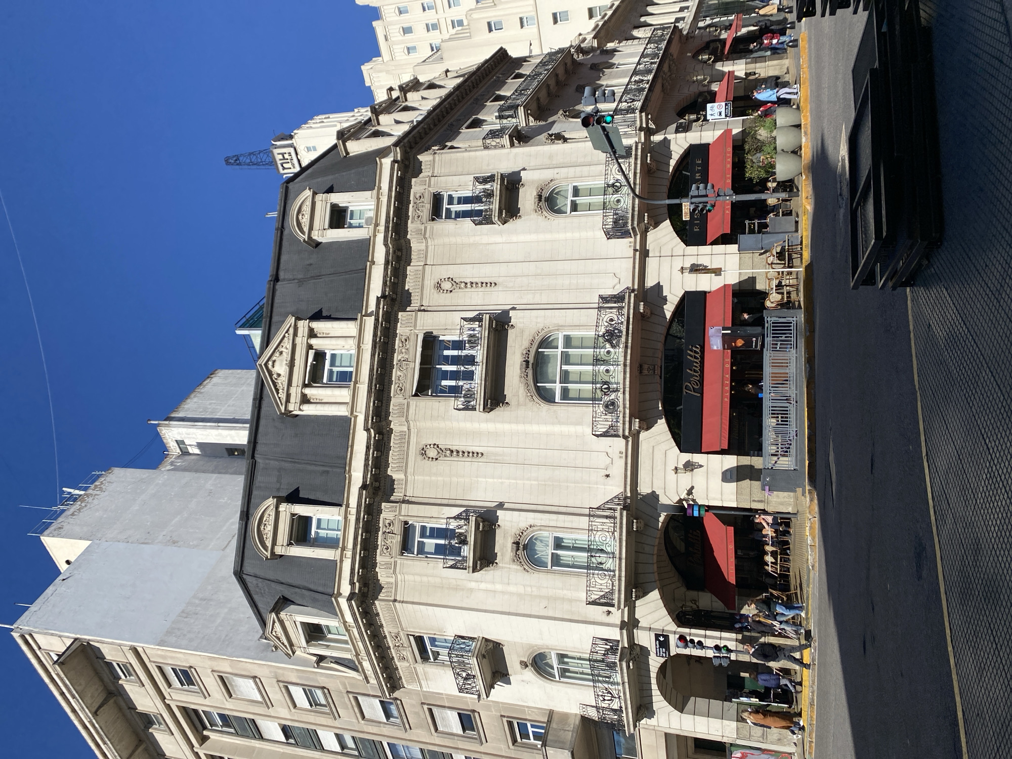 The image shows a building in Buenos Aires, Argentina. The building has a light-colored facade, dark mansard roof, and multiple levels with arched windows and wrought-iron balconies. A red awning covers a restaurant entrance labeled "Perutti, Plaza de Mayo." People are walking on the sidewalk and sitting at outdoor tables. A traffic light is visible. Across the street, a metal structure is visible. The sky is clear and blue.
