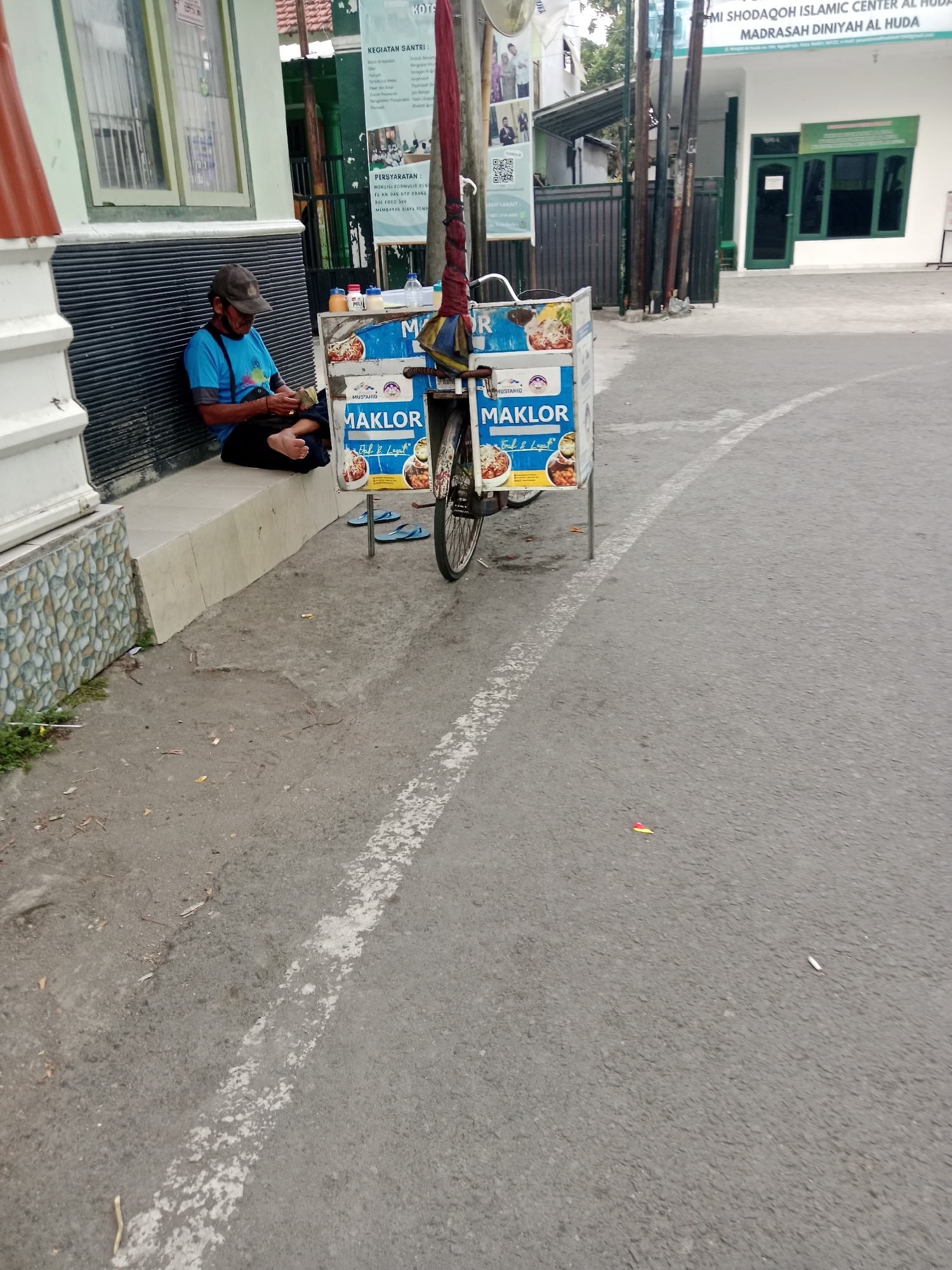 A man wearing a blue t-shirt and a cap sits on a low wall, facing a food cart. He is looking down and holding what appears to be money in his hands. The food cart, mounted on a bicycle, displays images of food and text that reads "MAKLOR." A red umbrella is attached to the cart. The scene is on a paved street, with a white line marking the edge. In the background, a building with green doors and windows is visible, along with signage that includes text in Arabic and Indonesian. The environment suggests an outdoor setting in Indonesia, likely a street or roadside.