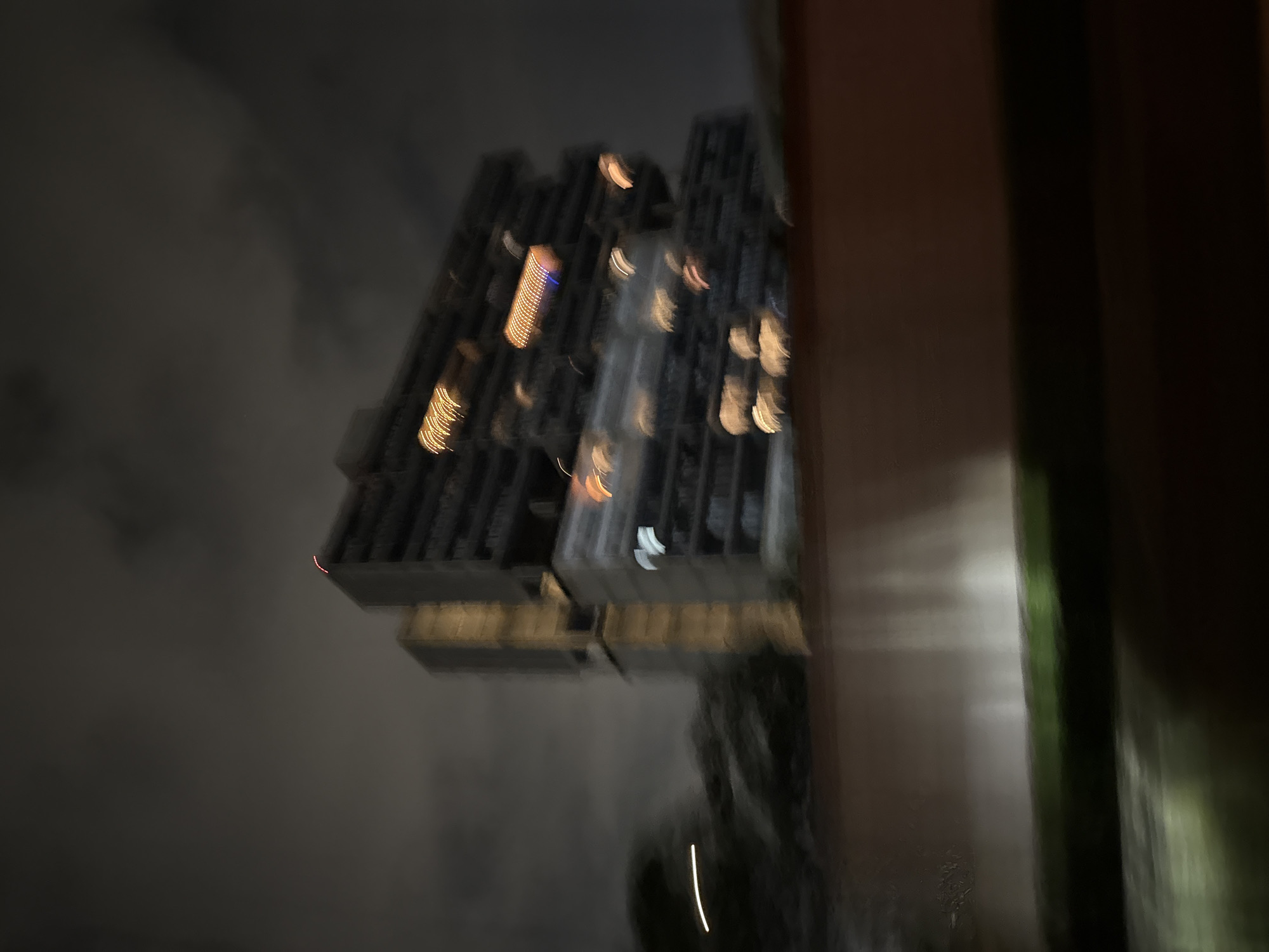 The image shows a tall building with numerous windows lit up at night. The building is dark gray with several illuminated sections, suggesting residential or office spaces. The surrounding environment appears cloudy and dark. There are elements of a structure or wall on the right side, which suggests the photo was taken from inside a building. There is no indication of the location being Santa Clara Ocoyucan, Mexico, or any specific identifiable landmarks or signs.