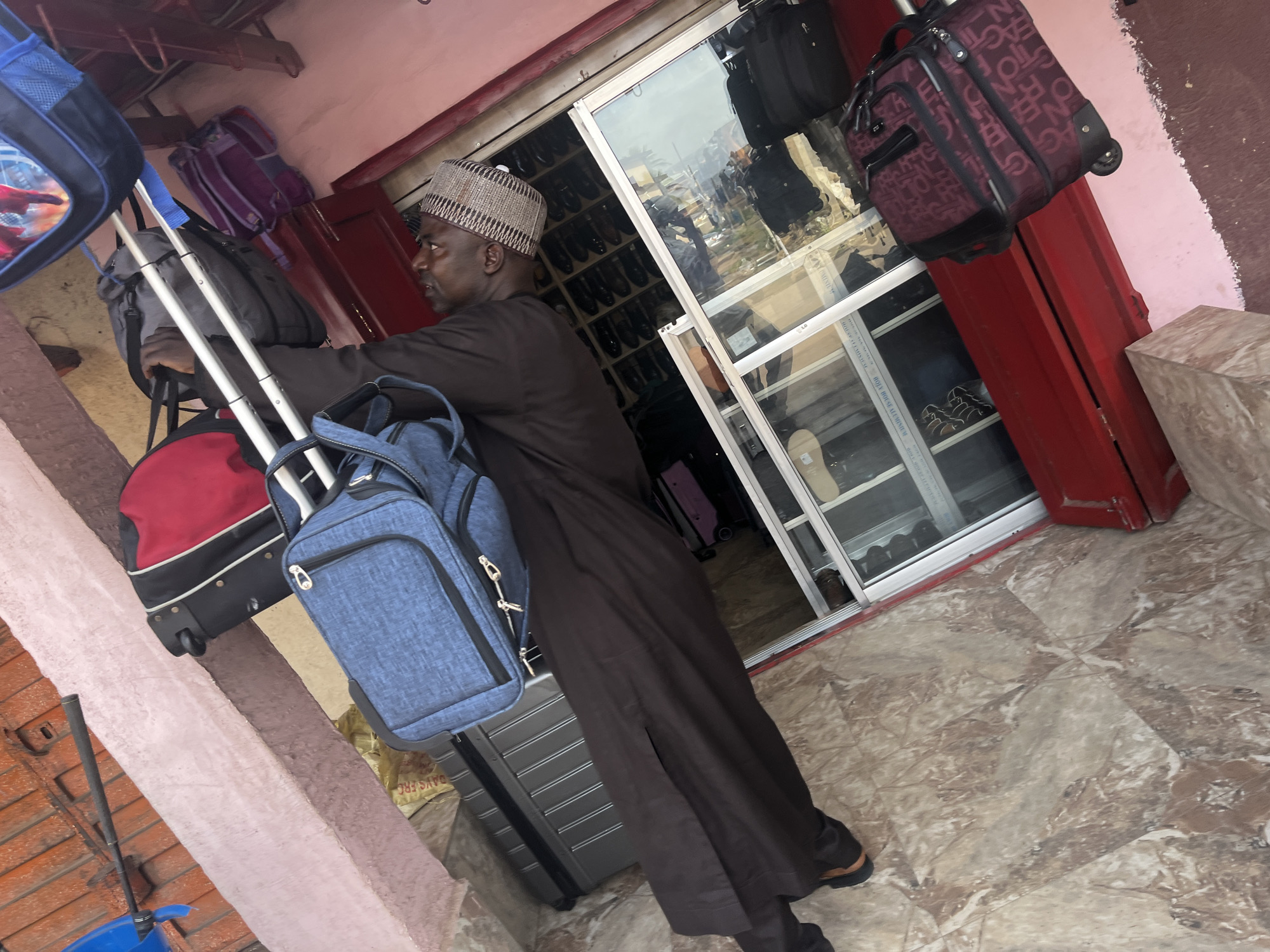 A man, appearing to be of African descent, is standing in front of a shop with luggage. He is wearing a dark brown traditional garment and a patterned cap. He is leaning against a rolling suitcase that has a blue duffel bag on top of it. To his left, there is another rolling suitcase with a red and black bag on top. More luggage, including a purple backpack, is visible behind him. The shop window displays various items, possibly clothing, and appears to be made of glass with metal framing. The floor is tiled with a marble pattern. The scene suggests a travel or transportation context, potentially at a bus station or a travel agency in Jos, Nigeria.