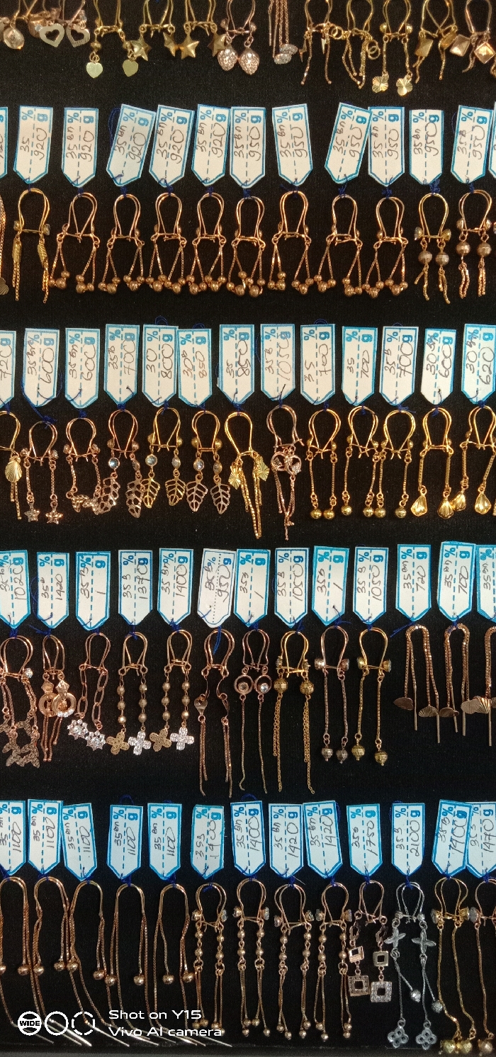 The image displays an array of earrings presented in a retail setting, likely within a jewelry store or stall in Maron, Indonesia. The earrings are arranged in approximately six horizontal rows against a uniform dark, non-reflective background, possibly black velvet or felt. The collection comprises numerous pairs of earrings, predominantly gold-toned, with some rose gold and silver-colored items. Each pair is individually showcased and features a small, white, rectangular price tag with a light blue border, attached by a thin string. Handwritten information on these tags includes percentages (e.g., "35 bn %," "30 %," "35.3 %"), a "g" suffix indicating grams, and three or four-digit numerical values representing weights or prices (e.g., "920," "600," "1400," "1750"). The earring designs are diverse, featuring simple hooks, elongated chains, multiple small beads, and various decorative elements such as hearts, stars, leaves, flowers, and geometric shapes. Some earrings are embellished with small, clear faceted stones. No individuals are present in the frame.