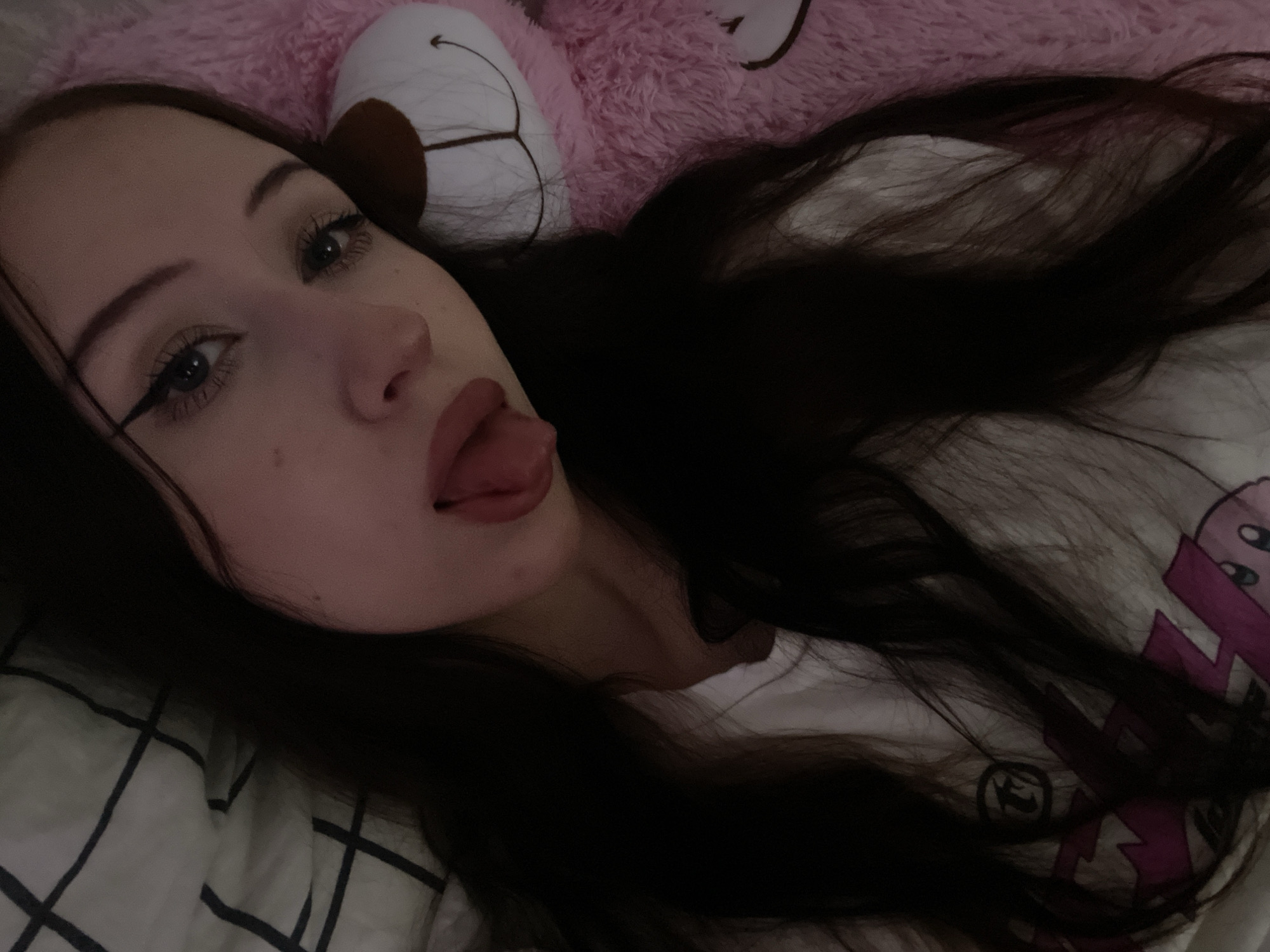 The image shows a young person with long, dark hair lying down, with their tongue sticking out. They are wearing a white t-shirt with a graphic design. To the right of the person is a large, pink plush toy, possibly a teddy bear. The background includes a white and black checkered pattern, likely from bedding. The lighting is dim, suggesting an indoor setting. There is no visible indication of the city of Votkinsk, Russia.