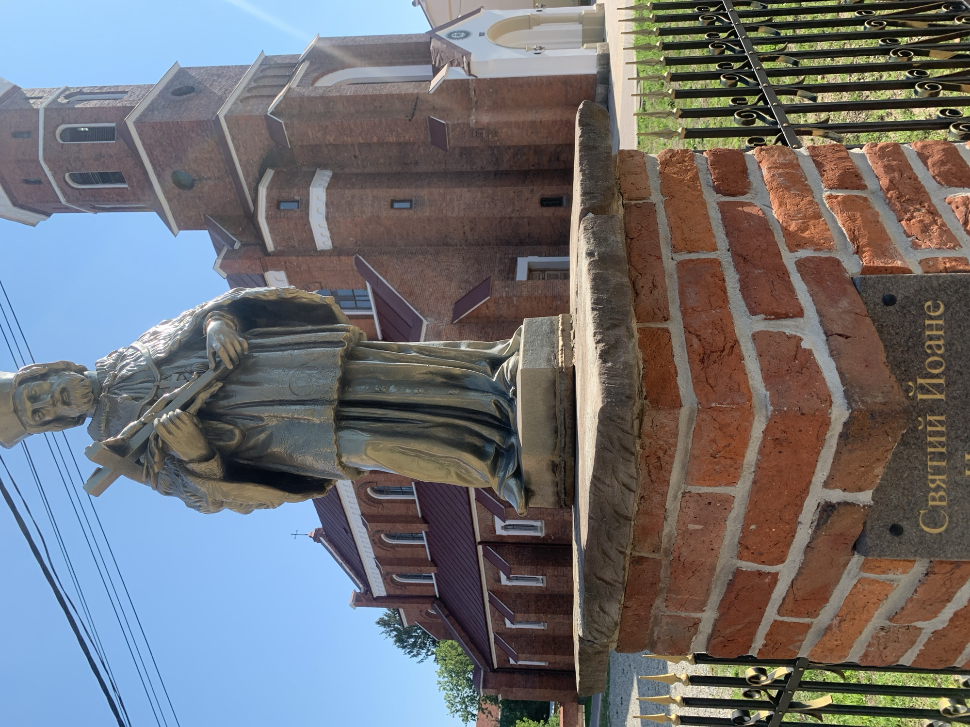 In Kolomyia, Ukraine, a bronze statue of a bearded man wearing a crown and holding a cross stands prominently in front of a red brick church. The statue is positioned on a brick pedestal with a stone top, adjacent to a decorative black metal fence with gold accents. The church features a tall, multi-tiered bell tower and arched windows, typical of religious architecture. The sky above is clear and blue, suggesting a sunny day. The inscription on a dark plaque at the base of the pedestal reads "Святий Йоане," which translates to "Saint John" in Ukrainian.