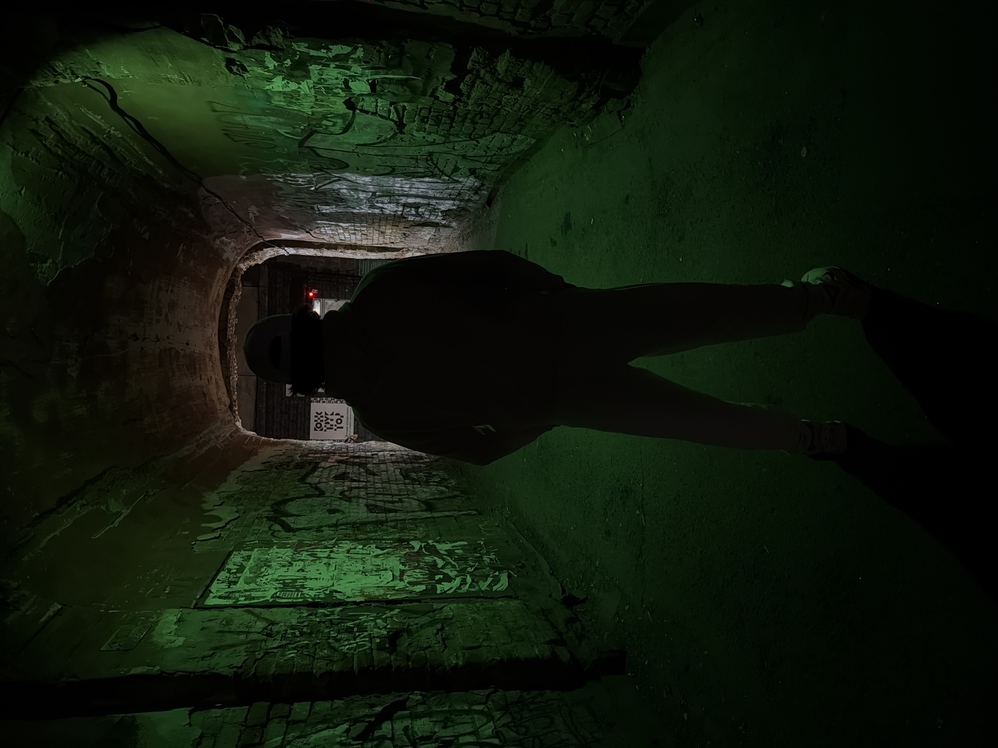 A person stands in the center of a graffiti-covered brick tunnel. The tunnel is illuminated by green light, with a red light visible further down the tunnel. The location is Krasnoyarsk, Russia.