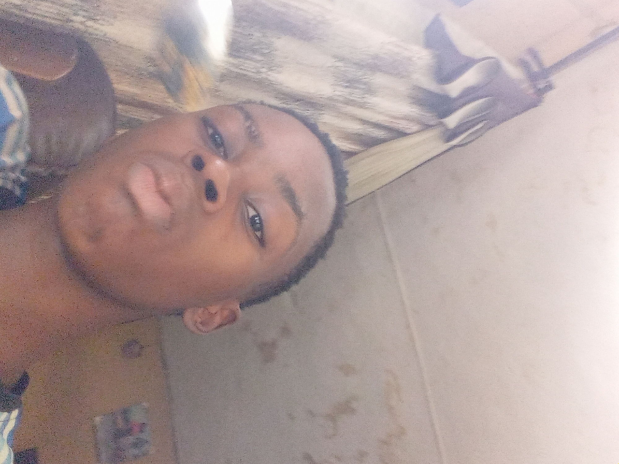 A selfie of a dark-skinned young man with short black hair, wearing a blue and white striped garment. He is looking directly at the camera with a slight pout. The background appears to be an interior room with beige walls. There is a striped curtain or cloth hanging in the upper right corner, and a blurry image or poster on the lower left side of the wall. The image is taken at an angle, with the man's face slightly off-center. There is no evidence to determine this image was taken in Onitsha, Nigeria.
