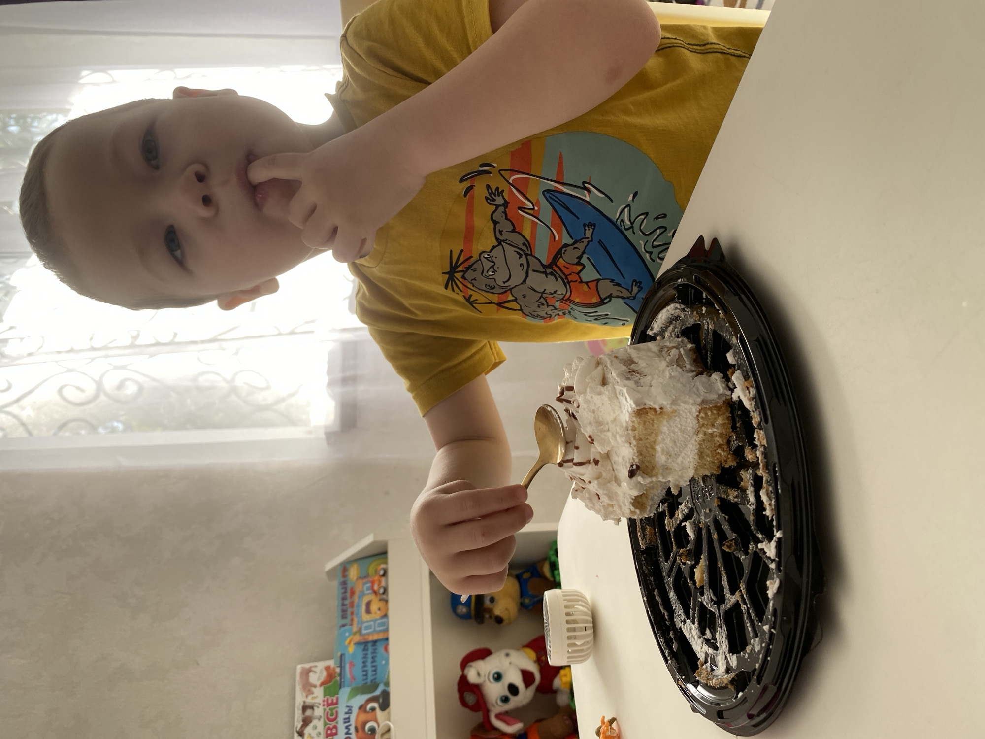 A child with fair skin and short hair is shown eating cake with a spoon. The child is wearing a yellow shirt with a cartoon graphic. A partly consumed cake with white frosting is present. Toys and books are visible in the background. The location in the city of Sochi, Russia is not possible to determine from this image.