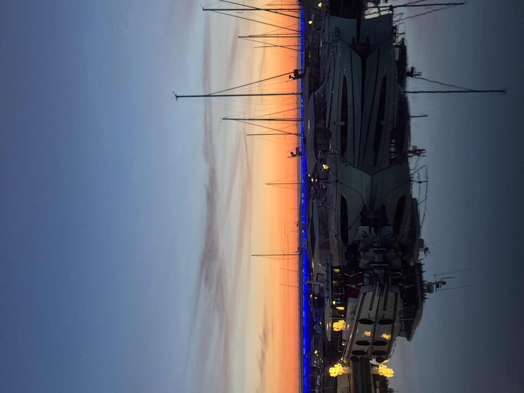 The image depicts a marina in Tallinn, Estonia, at dusk. Several yachts and sailboats are docked, their reflections shimmering in the calm water. The sky above transitions from a deep blue to a vibrant orange and pink hue as the sun sets. A line of bright blue lights runs along the waterfront, illuminating the scene. The yachts are various sizes, with some featuring tall masts and others appearing more like motorboats. The lighting suggests artificial lights from the boats and the waterfront are also contributing to the illumination. There are no people visible in the image.