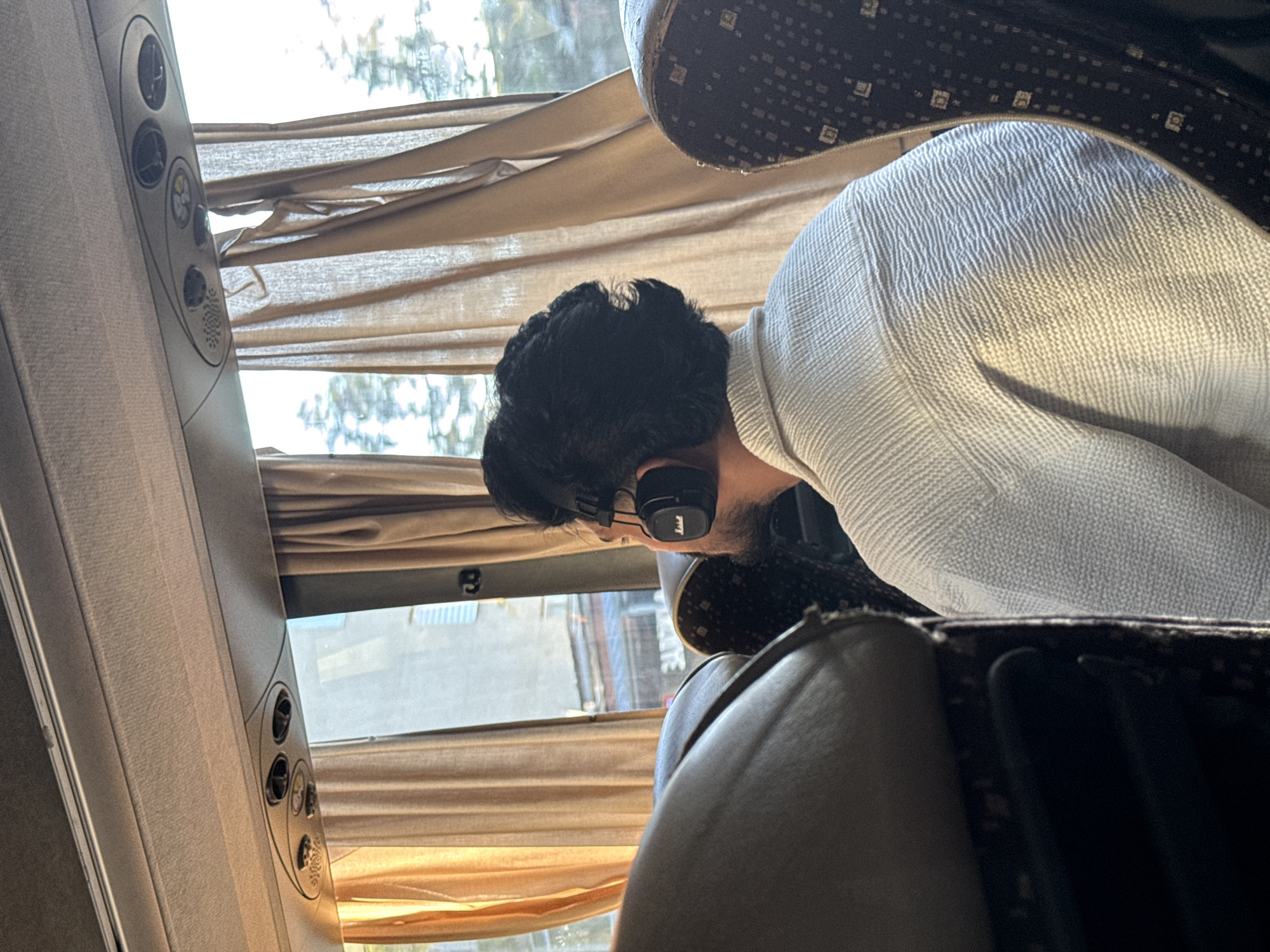 A person wearing black headphones is seated on a bus, facing away from the camera. The person has dark, curly hair and is wearing a light-colored textured shirt. The bus interior features brown curtains and seats with a patterned design. The background shows a view out the window of trees and buildings. The scene is captured indoors, likely during the daytime, with natural light filtering through the curtains.