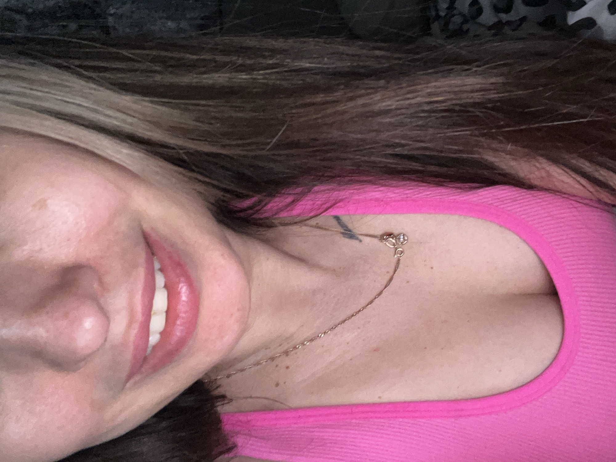 A close-up, tilted selfie depicts a fair-skinned woman with brown hair, smiling. She is wearing a bright pink top and a delicate gold chain necklace. A tattoo is visible on her chest. The background is blurry and partially obscured by the woman's hair, revealing a dark, patterned surface. The location and city are not specified.