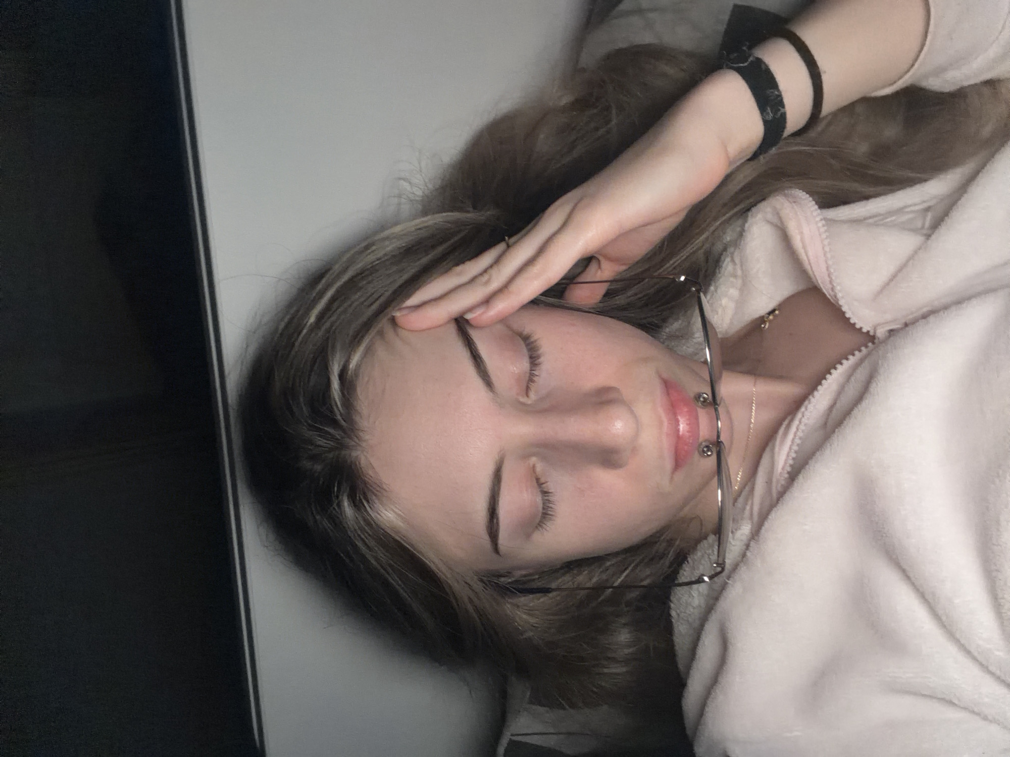A young woman lies on her side, eyes closed, her right hand resting on her forehead. She is wearing a light pink zip-up jacket and two black wristbands on her left wrist. Her glasses are askew, resting against her face and clothing. The background is out of focus, but a portion of a dark surface, possibly a piece of furniture or wall, is visible to the left. There's no information that suggests the image was taken in the city of Plovdiv, Bulgaria.