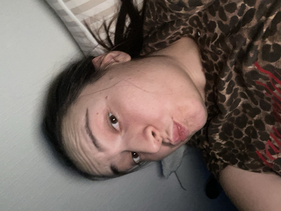 A woman lies on her side, gazing into the camera with pursed lips. She wears a brown and black leopard print top with red accents. Her dark hair is pulled back, revealing her forehead and face. The background includes a white pillow with faint gray stripes and a plain light gray wall. The lighting suggests an indoor setting, possibly a bedroom. The woman's expression hints at a playful or teasing mood.