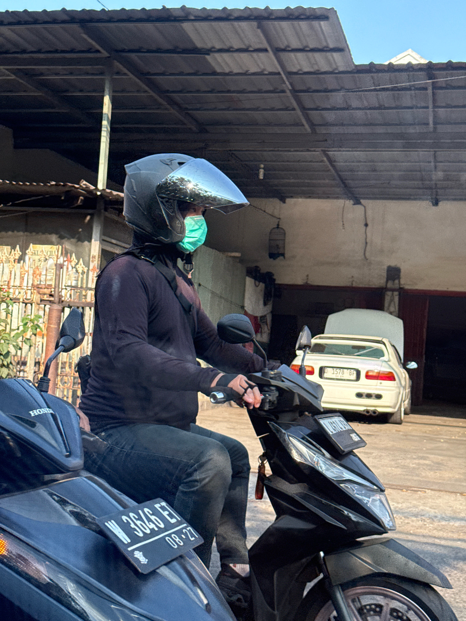 A person wearing a black long-sleeved shirt, blue jeans, a black helmet with a reflective visor, and a green face mask is seated on a black scooter in an outdoor setting. The scooter has a license plate that reads "W 3646 ET 08-27". In the background, a white car with its hood open is parked in front of a building that appears to be a garage or workshop. The environment suggests a sunny day in a somewhat industrial or residential area, potentially in Surabaya, Indonesia, given the presence of common Indonesian license plate formats and architectural styles. The person is looking to the right, possibly waiting or about to depart. The scene is captured from a slightly low angle, emphasizing the scooter and its rider.