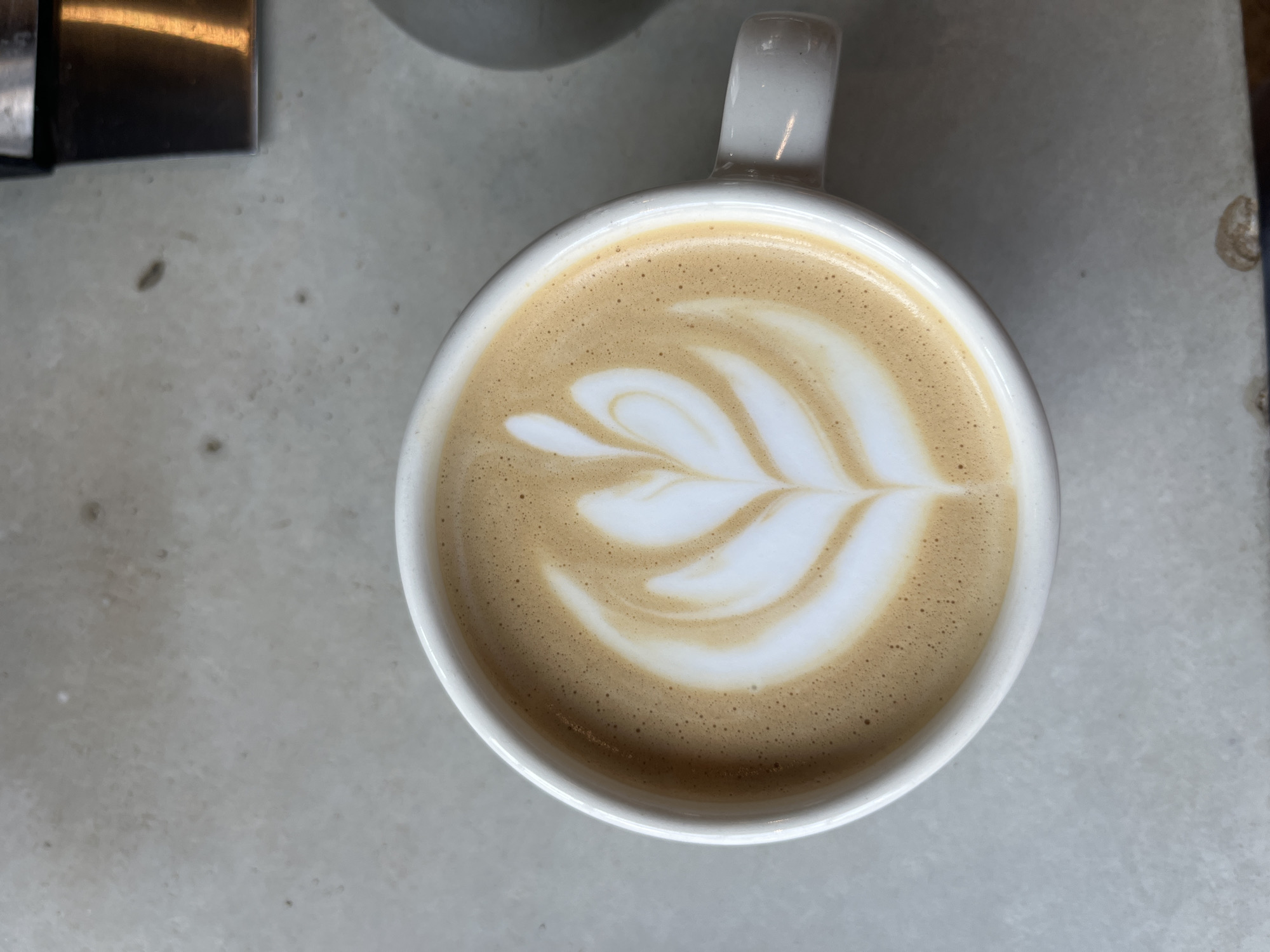 An overhead shot captures a white ceramic cup filled with latte art. The latte art features a symmetrical pattern of white foam resembling a tulip or a swan's neck against the backdrop of the coffee's creamy brown surface. The cup sits on a light-colored, textured surface, possibly a countertop or table. To the right of the cup, a sliver of a dark gray, rounded object is visible, and further to the upper right, a portion of a metallic appliance with a glowing orange light can be seen. The scene appears to be an indoor setting, likely a cafe or kitchen, in Kaliningrad, Russia. There are no people visible in the image.