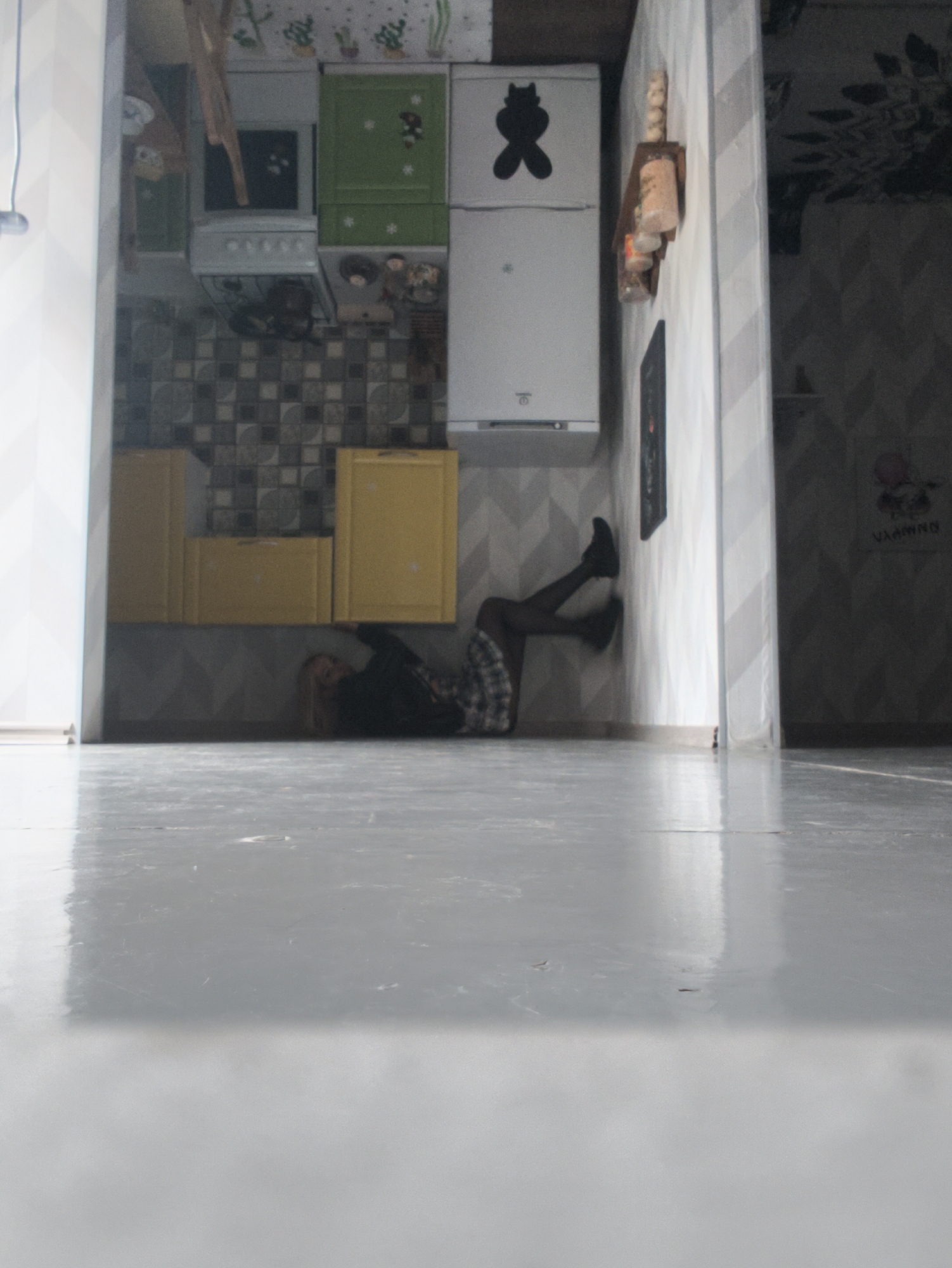 The image captures an inverted domestic scene, creating an optical illusion. A person, appearing to be a woman, is positioned as if lying on a wall, but due to the inverted perspective, she seems to be on the floor or floating in an upside-down room. She wears a dark top, a plaid skirt, black tights, and black shoes, with her legs bent and feet pressed against the vertical surface.
The setting is a kitchen connected to another room or hallway. The kitchen features white and green upper cabinets, a white stove, and yellow lower cabinets. A white refrigerator, adorned with a black cat silhouette sticker, stands against a wall. Above it, a wooden spice rack with glass jars is mounted. The kitchen area is tiled with dark, square-patterned tiles, while other walls display a light-colored chevron pattern wallpaper.
The foreground is dominated by a highly reflective, glossy grey floor surface, mirroring light and showing minor imperfections. In the adjacent room, visible through an archway, there is a darker patterned wallpaper and faint text on the wall that appears to read "VIAMINA".
The overall image is bright, suggesting daylight, with the reflective floor enhancing the sense of space and illusion. The scene evokes a playful and surreal atmosphere. The location context is Sevastopol, Ukraine.