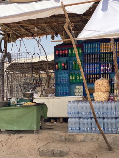 The image depicts an outdoor shop in Sard-E 'olya, Afghanistan. A weathered white structure with blue shelves displays beverages including Coca-Cola, Sprite, water bottles, and assorted canned drinks. Stacks of bottled water sit below the shelves. To the left, a scale and a tray of food, possibly grains, are on a green-covered table. A rudimentary fence surrounds the area. Two makeshift canopies provide shade. A wooden post acts as a support for the structure. The ground is dusty, with concrete steps leading up to the shop.
