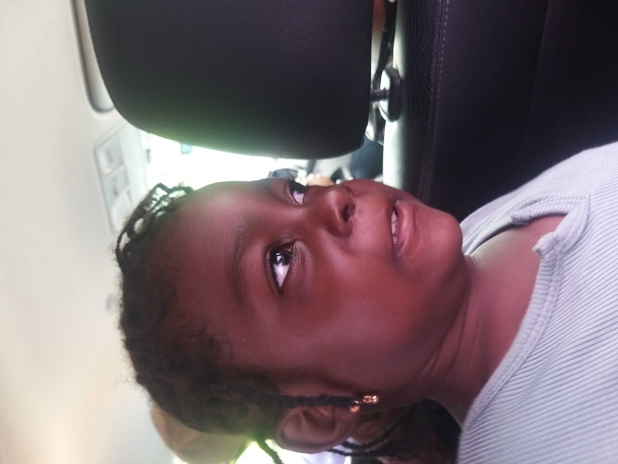In this close-up, vertically oriented photograph, a young Black girl is the sole focus, her face filling most of the frame. She is looking upwards and to her right with a slight, closed-mouth smile. Her dark hair is styled in braids, some of which frame her face. She wears a small, gold hoop earring in her left ear. Her skin is a deep brown, and the lighting creates a warm, reddish glow on her face. She is wearing a light blue, ribbed shirt.
The girl appears to be in the back seat of a car, with the dark headrest of a car seat visible behind her. A portion of a car's interior, including what looks like a dome light, is visible in the upper left quadrant of the image. The background is blurred, suggesting movement or a shallow depth of field, but the overall impression is that of being inside a vehicle. No specific time of day or weather conditions are discernible, though the lighting suggests daylight. The context provided indicates the location is Dukku, Nigeria. No text is visible in the image.