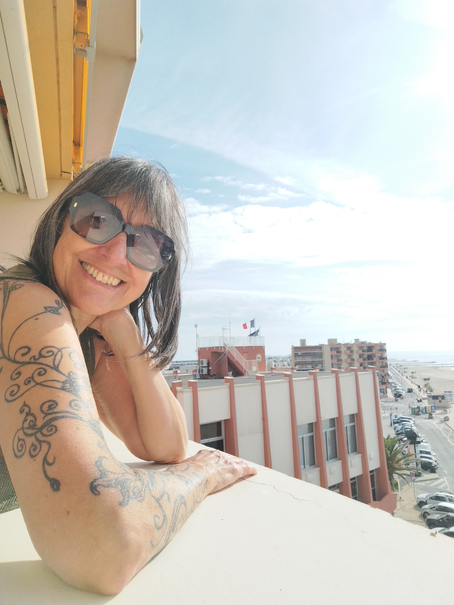 The image is a medium shot of a woman with shoulder-length gray hair and dark sunglasses smiling at the camera from a balcony. Her left arm is extensively tattooed with swirling, floral-like designs. She is leaning on the balcony railing with her arm bent and her hand near her cheek.
Behind her is a cityscape. There are several buildings, including a white and salmon-colored building directly behind the railing. In the distance, there is a beach and the sea under a partly cloudy sky. There are parked cars on the street and a palm tree visible. Based on architectural features, the presence of a beach and cars, and the flags, the location is Palavas-les-Flots, France.