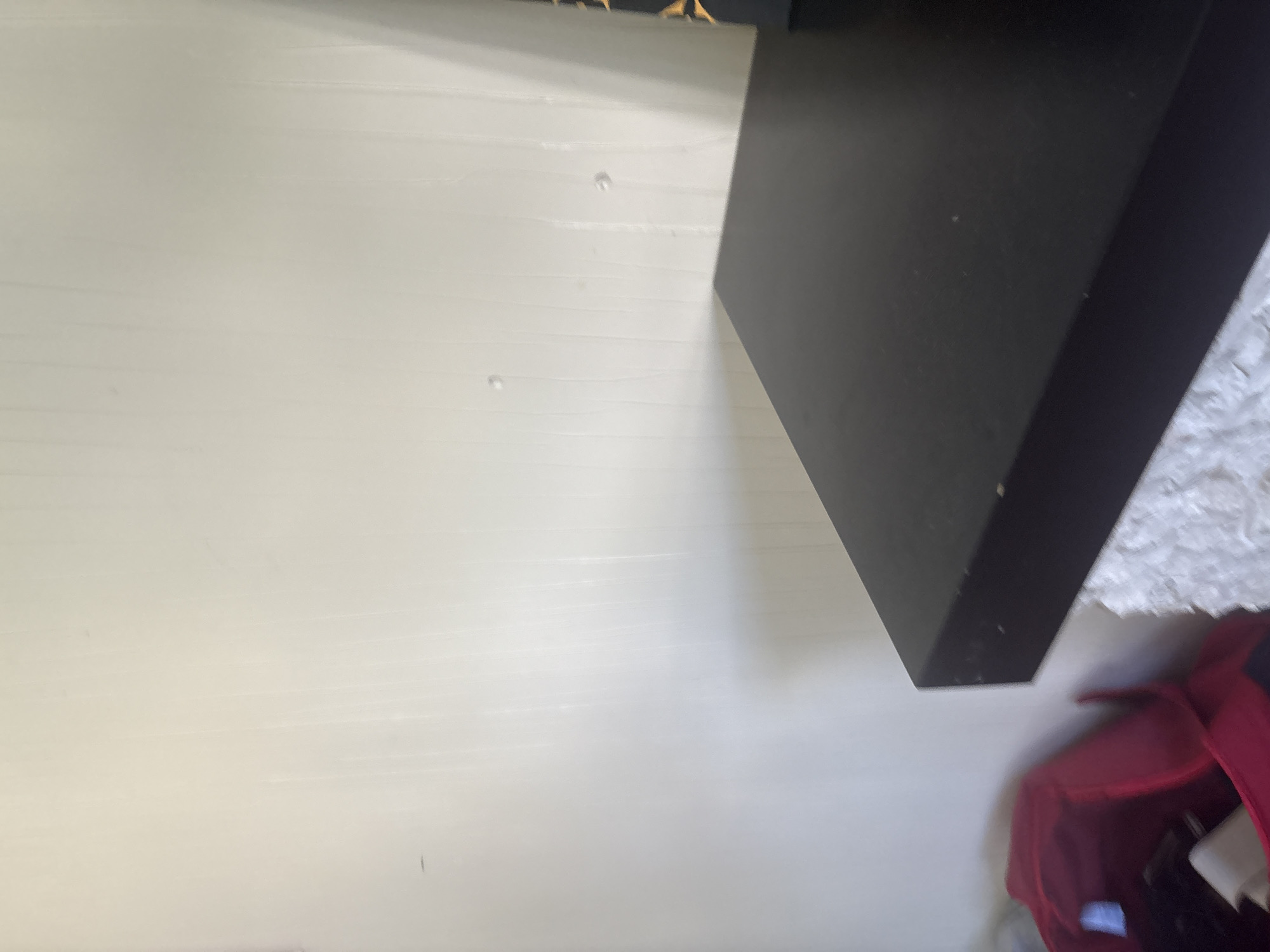 The image shows a corner of a room with a black shelf or table in the foreground. The wall behind is light-colored and appears to have some vertical texture. A red bag is visible to the left of the shelf. There are no people or any other discernible objects in the scene. The location is not determinable from the image content.