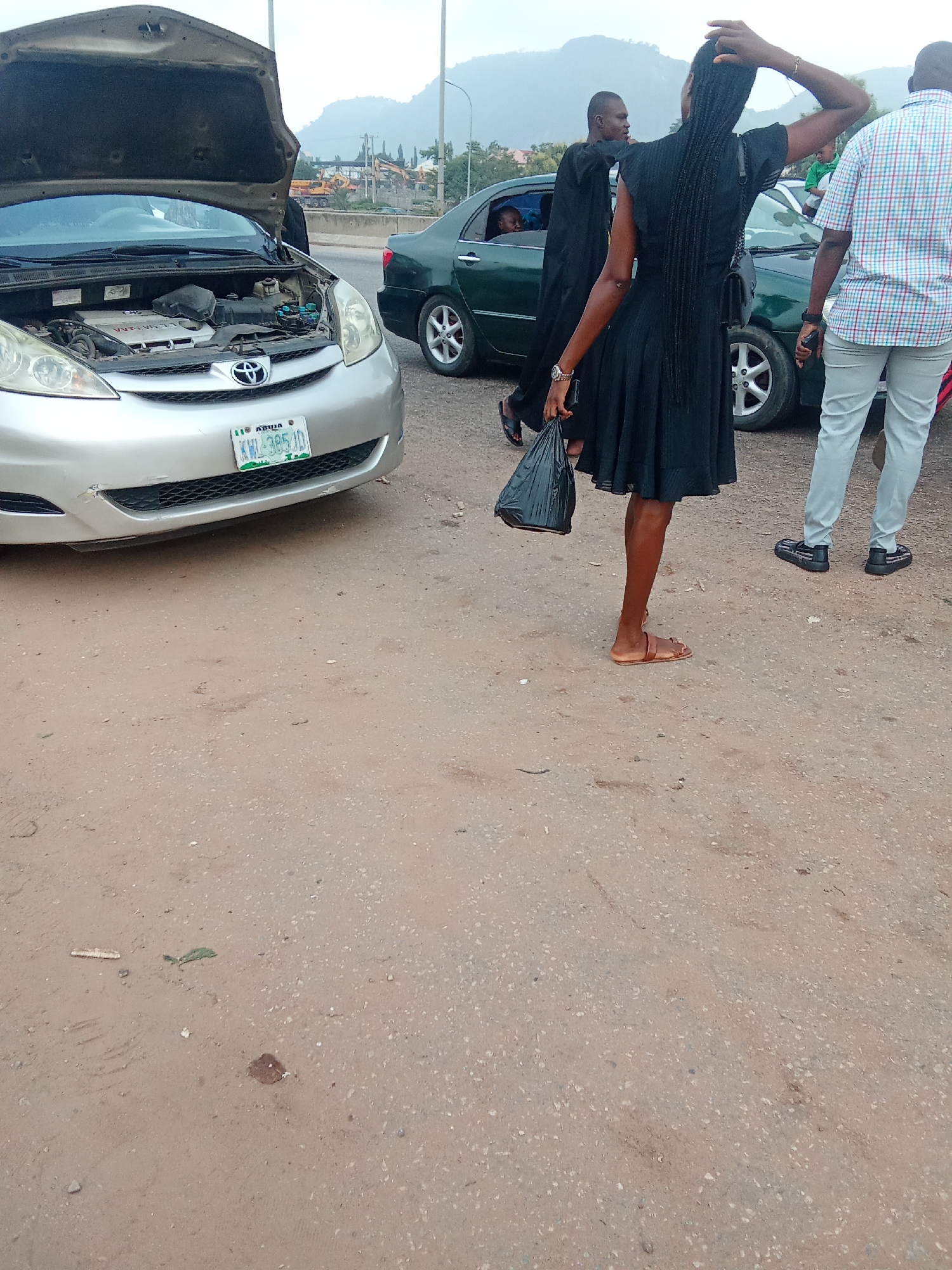 In Kagini, Nigeria, on an unpaved road, a silver Toyota Sienna minivan with its hood open is parked. The minivan's engine is visible. A dark-colored sedan is parked behind the minivan, and a green sedan is to the right of the minivan. A man in a checkered shirt and grey pants stands to the right of the scene. A woman in a black dress with braided hair is standing between the two sedans, holding a black plastic bag. Her left hand is on her head, and her right hand is by her side. Another man in a black robe is partially visible in the background, looking towards the woman. The environment is dusty with sparse vegetation and a mountainous backdrop.