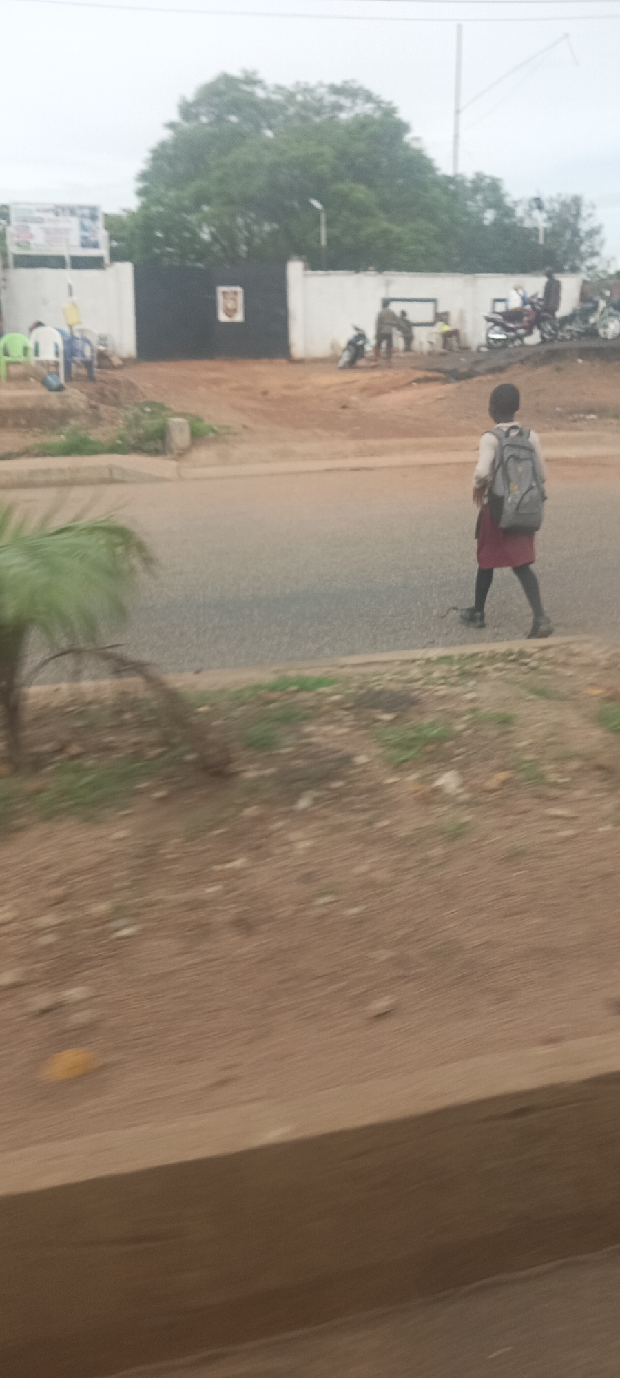 A blurred, low-resolution image taken from a moving vehicle captures a street scene with a school-aged child walking across the road. The child, dressed in a white shirt and a maroon skirt with black stockings, carries a gray backpack. In the background, a white wall with a black gate is visible, possibly marking the entrance to a school or compound. Several motorcycles are parked near the gate, and a few people are standing around. Green and blue plastic chairs are also present. The environment appears to be an urban setting with a paved road and dirt/gravel areas on the sides. The sky is overcast. The scene is consistent with a location in Jos, Nigeria, given the architecture and general ambiance.