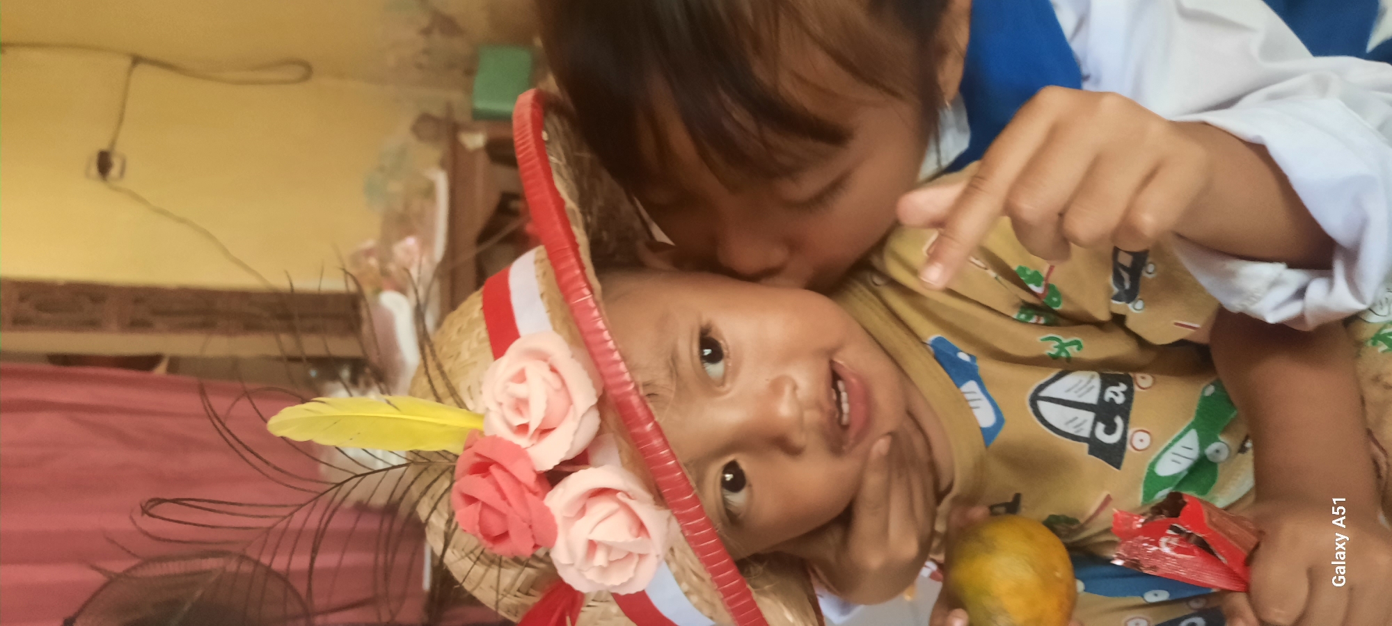 In Ploso, Indonesia, a close-up shot captures a young child wearing a straw hat adorned with red and pink fabric roses and a yellow feather. The child has dark eyes and is looking slightly to the right with their mouth open. Another child is gently kissing the cheek of the child in the hat, partially obscuring their face. The child in the hat holds an orange in their left hand and a red candy wrapper in their right. They are wearing a yellow t-shirt with a pattern of cartoon vehicles and the letters "Ca". The background is a softly lit room with a pink curtain on the left and a yellow wall with an electrical outlet and cord on the right. The lighting suggests an indoor setting, possibly a home.