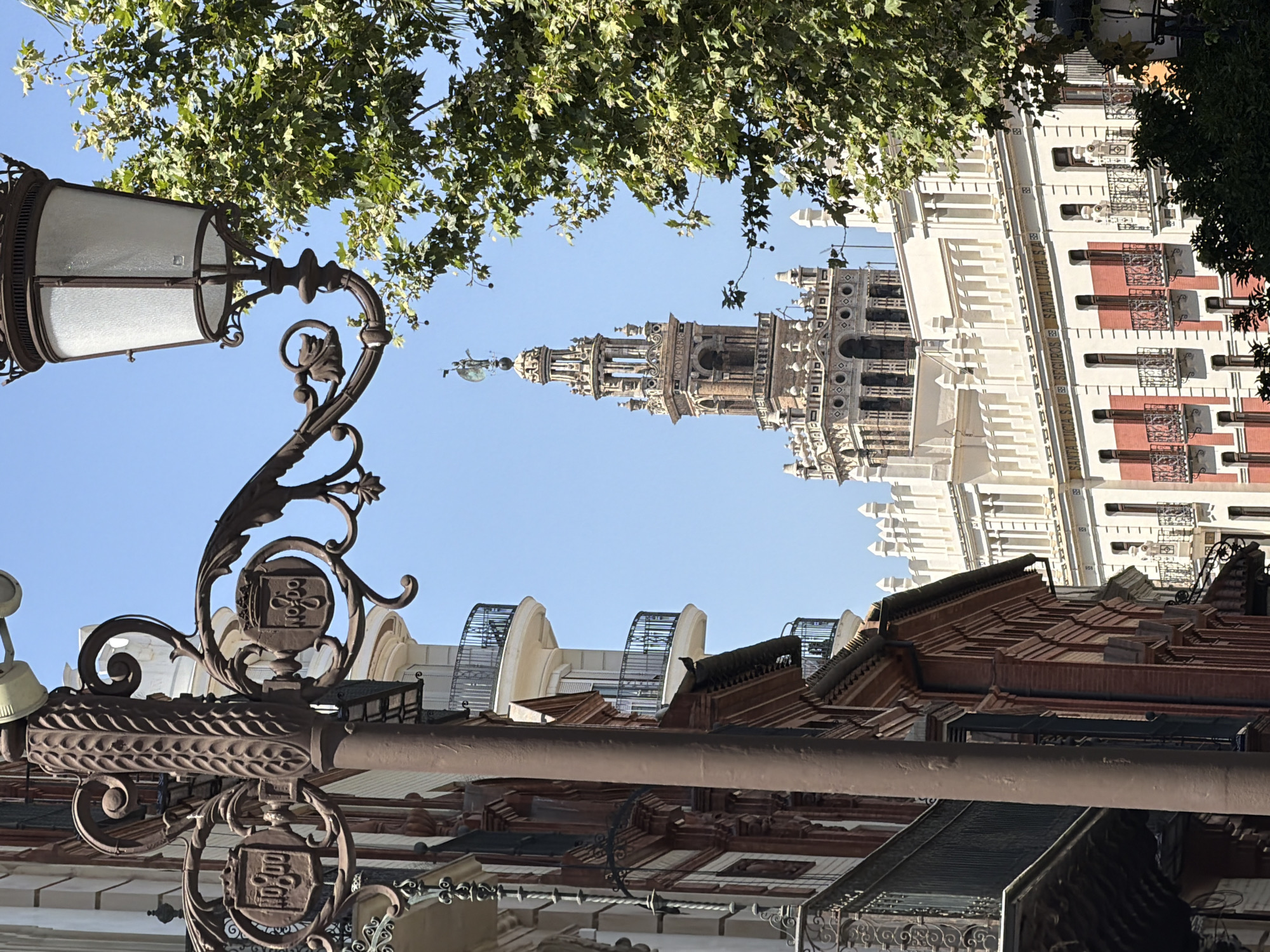 The image captures a street scene in Seville, Spain, with a prominent ornate lamppost in the foreground. Behind the lamppost, a tall, intricately designed tower, likely the Giralda tower, rises against a clear blue sky. The tower is flanked by buildings with traditional Spanish architecture, featuring balconies with wrought-iron railings and decorative facades. Green foliage from trees partially obscures the view of the buildings and the tower. The scene conveys a sense of historic European urban landscape on a sunny day.