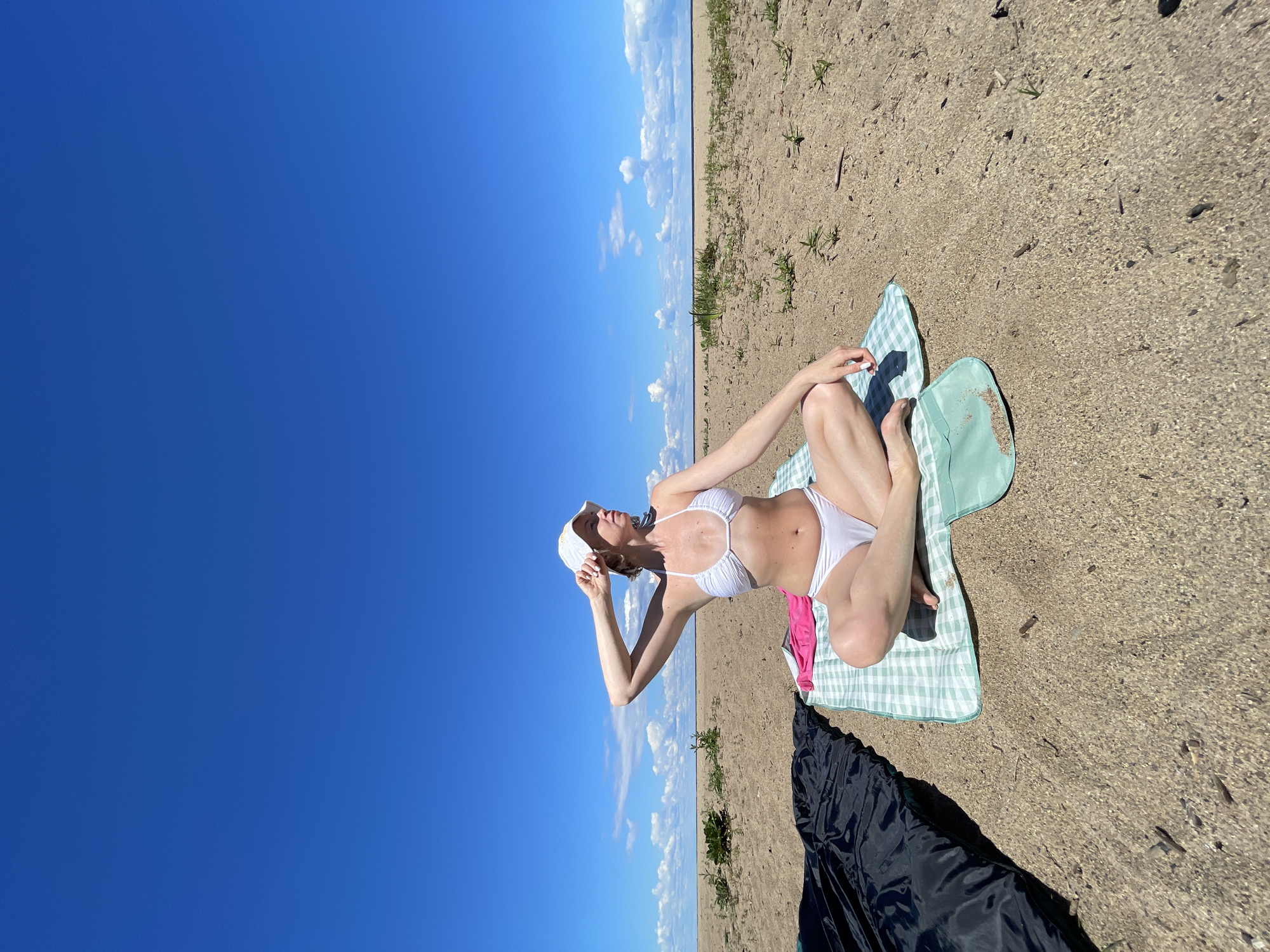 A woman in a white bikini and a white cap sits on a checkered blanket on a sandy beach. She is leaning back with her legs crossed and her arm resting on her knee, looking up at the sky. A pink towel is tucked under her leg. To the left of the frame, a vast expanse of bright blue sky dominates, with a thin strip of clouds on the horizon. The beach is sandy with sparse patches of green vegetation. A black tarp is partially visible in the foreground. The scene appears to be set on a sunny day, likely during daylight hours. The overall mood is one of relaxation and enjoyment of the outdoors. The location is identified as Belozersk, Russia. No visible text is present in the image.