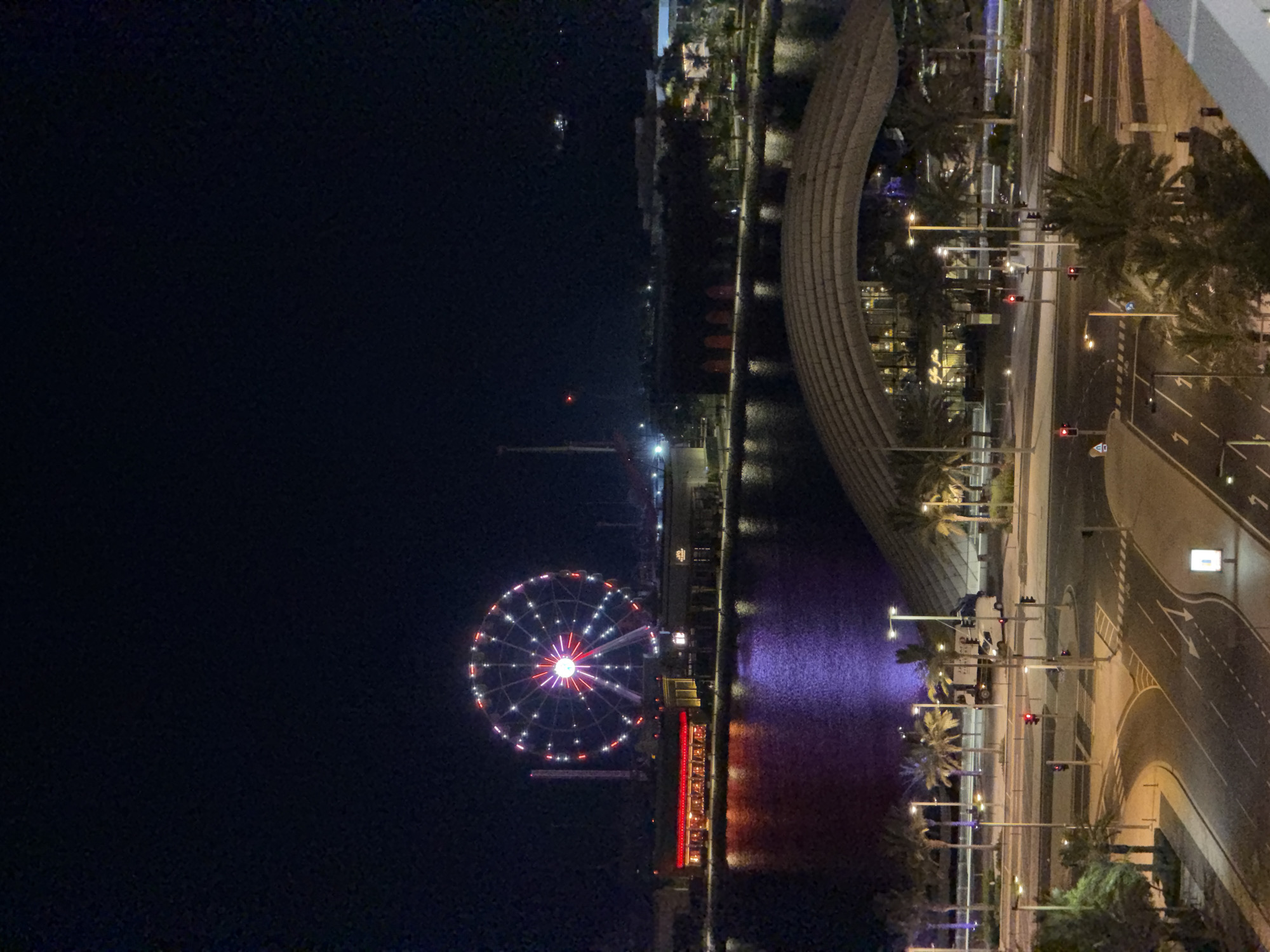 This is a nighttime aerial photograph of a waterfront entertainment area. The most prominent feature is a large Ferris wheel, illuminated with red and white lights, standing tall against the dark sky. In front of the Ferris wheel, a body of water reflects the lights of the surrounding buildings, which appear to be restaurants and entertainment venues. The water has a purplish hue. Further in the foreground, a modern building with a distinctive arched roof is visible. Below this, a street intersection with traffic lights and palm trees is illuminated by streetlights. Several vehicles, including a truck and a white van, are present on the road. The scene depicts a lively urban nightscape, likely a tourist or leisure destination.