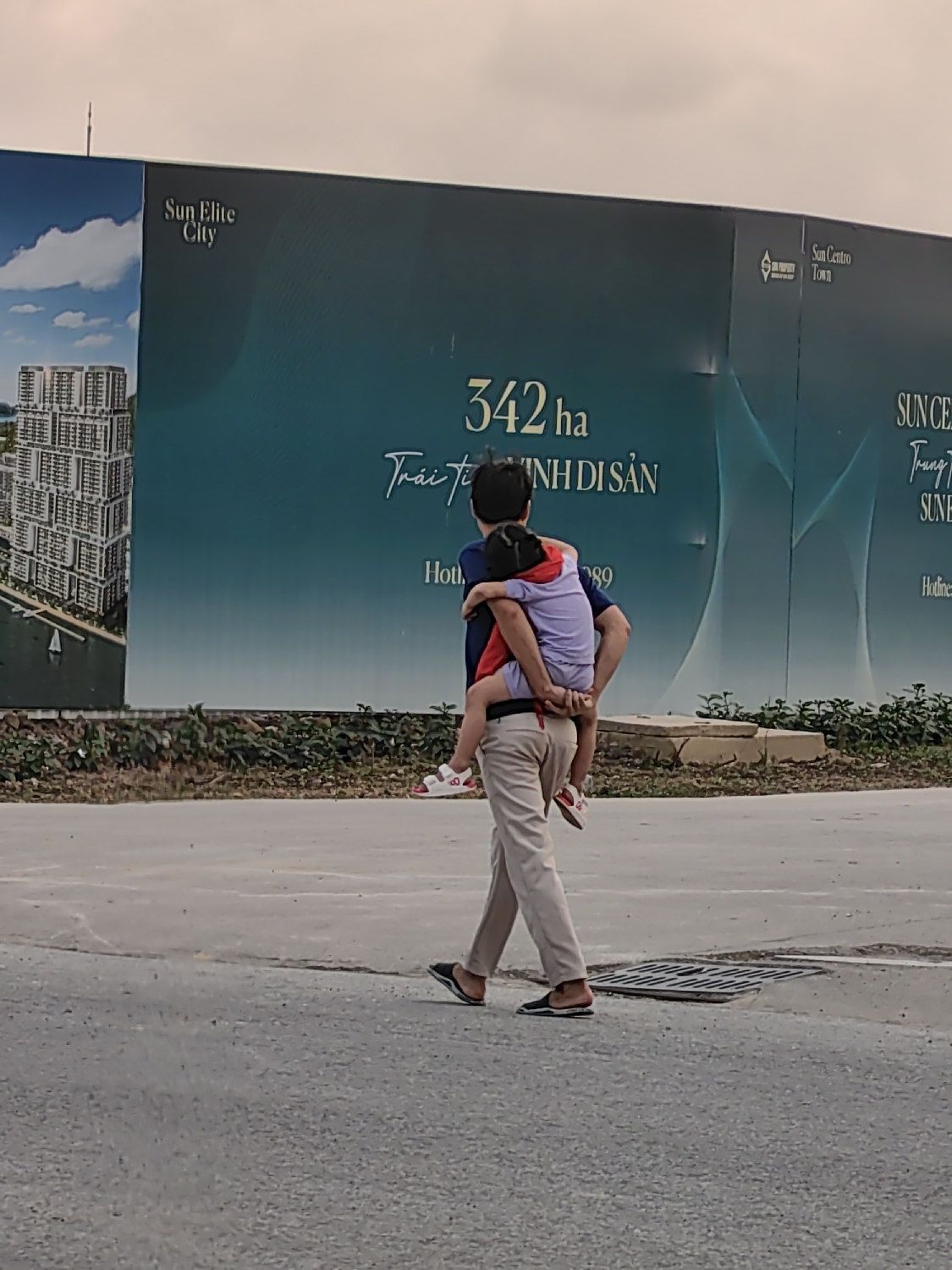An adult, likely male, is depicted walking away from the camera, carrying a child piggyback on a paved road. The adult wears a dark blue shirt, light beige trousers, and dark slippers. The child, dressed in a light purple top and red shorts with white sandals, clings to the adult's back.
The background features a large, dark blue-green billboard advertising a real estate development. On the left panel, "Sun Elite City" is visible above an illustration of high-rise buildings. The central section displays large white text: "342 ha" (hectares) and "Trái tim DI SẢN," meaning "Heart of Heritage." A partial "Hotline" number, ending in "89," is also visible. Further right, the billboard shows "Sun Centro Town," with additional fragmented text "SUN C" and "SUN E."
The scene is set outdoors during the daytime, under an overcast sky. Sparse greenery is present at the base of the billboard, and a manhole cover is on the right side of the road. The visible Vietnamese text and real estate advertisements suggest the location is a developing area in Bai Chay Ward, Vietnam. The image captures a quiet moment of familial care as the adult carries the child.