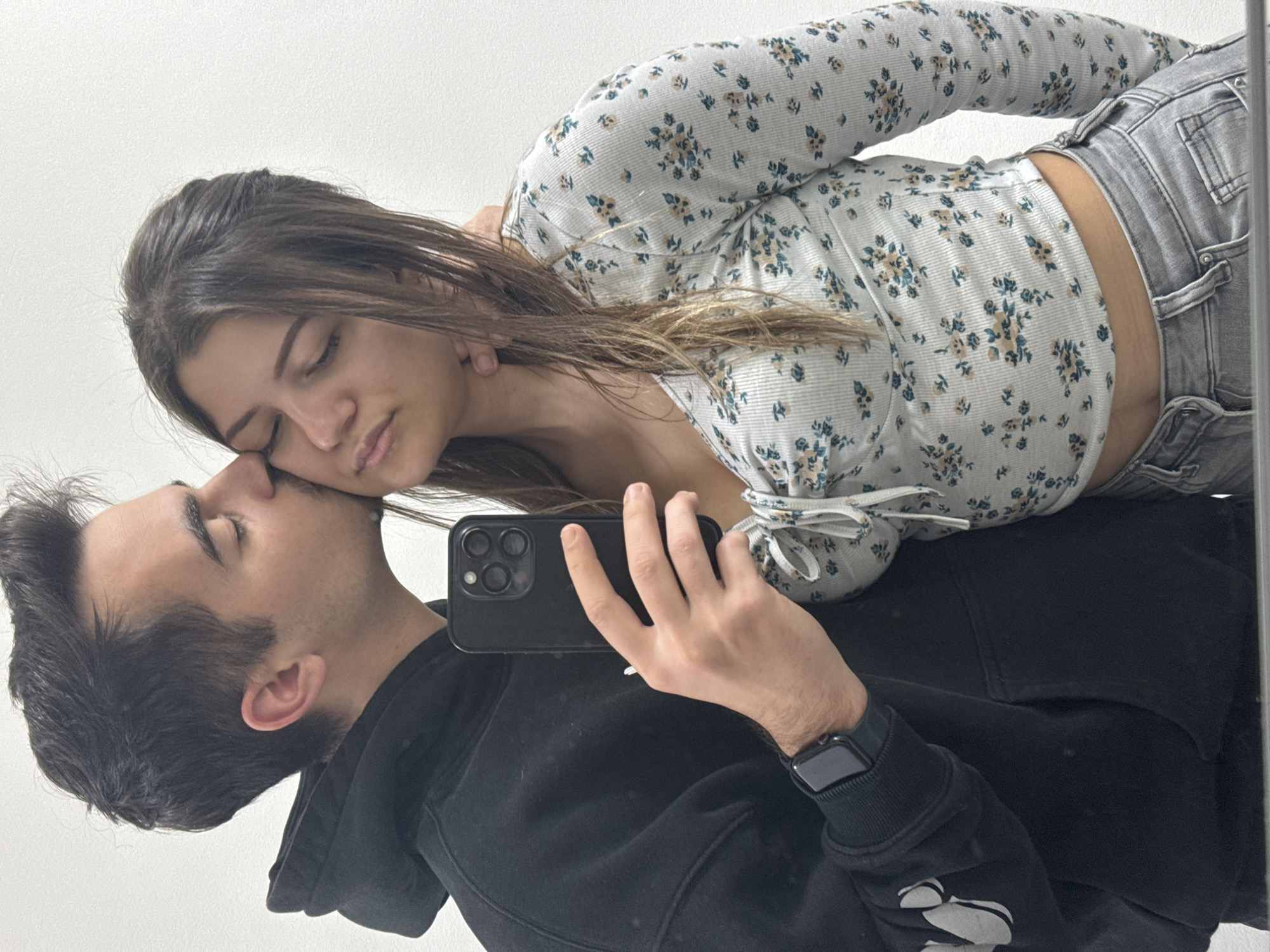 A couple is posed in front of a mirror, likely taking a selfie. The man, wearing a black hoodie and a smartwatch, holds a black smartphone. He is kissing the woman on the forehead. The woman, with brown hair, wears a floral long-sleeved shirt and grey jeans. The location is difficult to ascertain, but based on the subject's apparent geolocation it is likely Sofia, Bulgaria. The background appears to be a plain white wall.